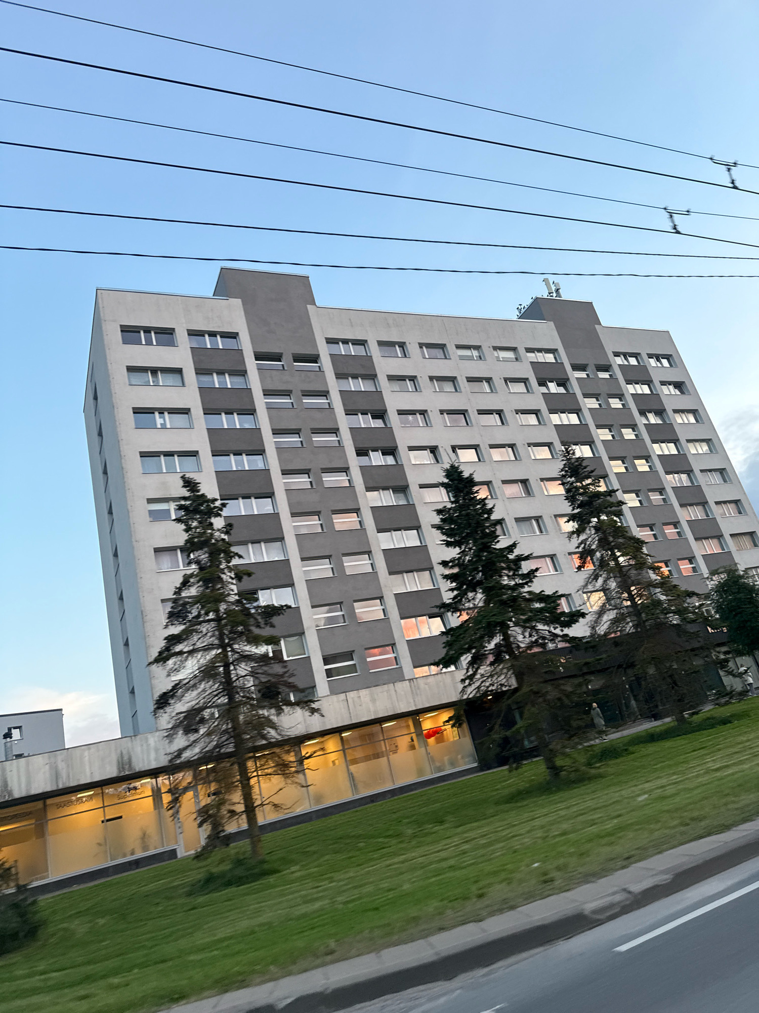 The image shows a tall, multi-story apartment building in Tallinn, Estonia, under a clear sky with overhead wires. The building is predominantly white with dark gray vertical sections and horizontal bands. Numerous windows, some illuminated with warm light, are visible across the facade. In front of the building, there is a grassy area and several tall, slender evergreen trees. A lower section of the building, possibly commercial space, has large windows with frosted glass and illuminated interiors. A street with a curb and road markings is visible in the foreground, suggesting the photo was taken from a vehicle or roadside.