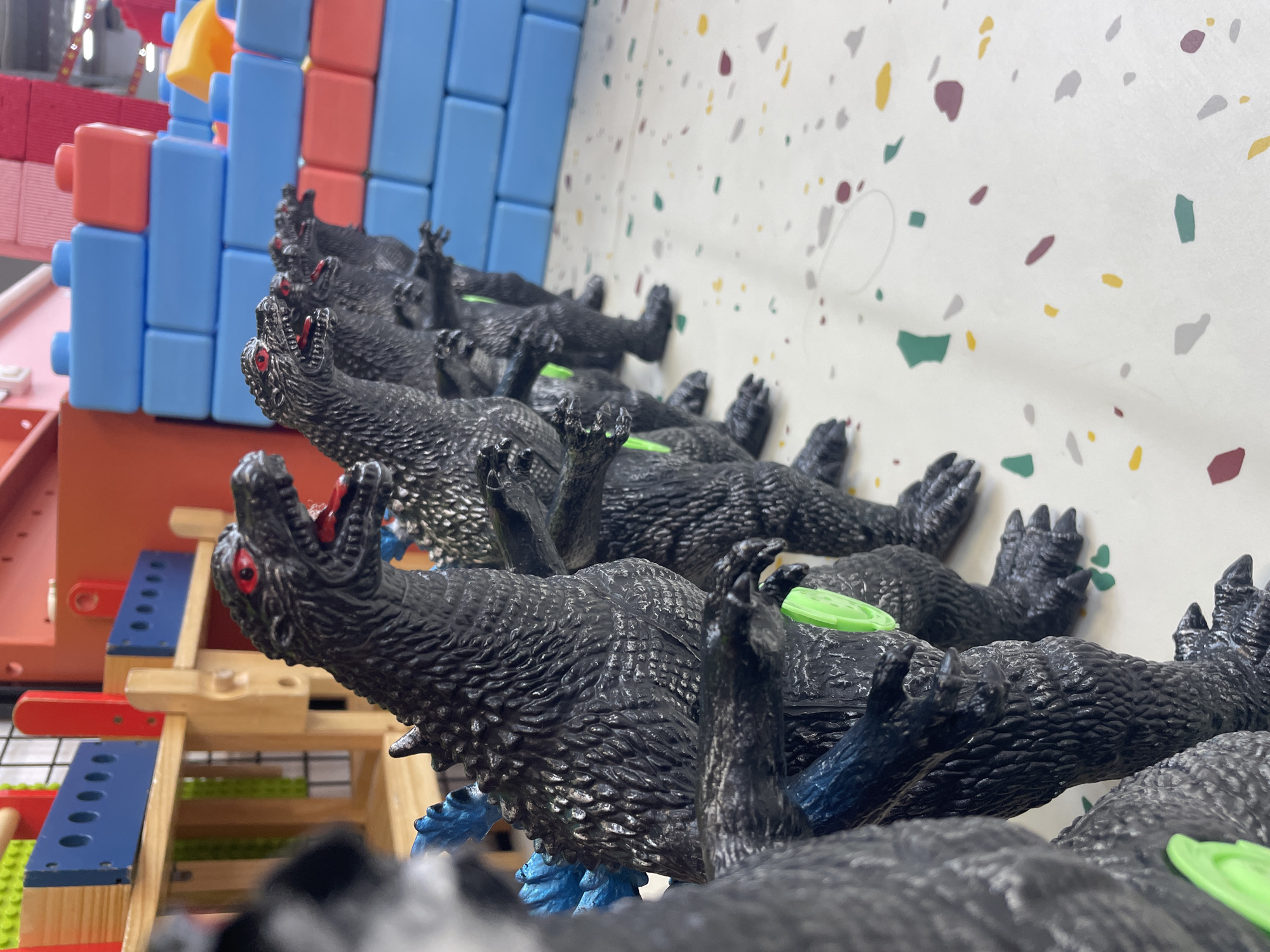 The image shows a row of black toy dinosaurs, some with open red mouths, arranged in a line against a white wall with multicolored speckled pattern. To the left of the dinosaurs, there are colorful building blocks and a wooden structure with red and blue accents. The location is identified as Nha Trang Ward, Vietnam.