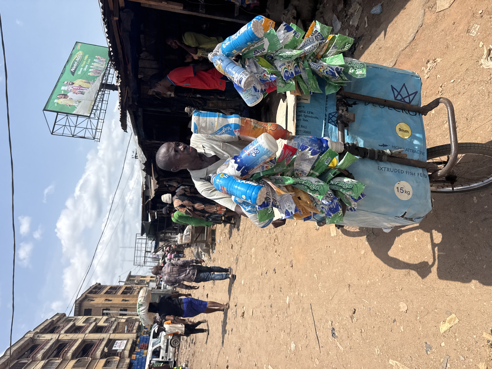 A man pushes a heavily loaded tricycle cart through a bustling street in Onitsha, Nigeria, under bright daylight. The scene depicts a vibrant market or commercial area with unpaved ground and buildings lining the thoroughfare. The man, visible in a light-colored shirt, is focused on maneuvering his cart.
The tricycle cart is laden with numerous items, including stacks of plastic bottles, primarily yogurt or milk drinks like "Nutri-Milk," "Diva," "Yoghu," and "Apple." Additionally, many small, brightly colored pouches, possibly snacks or drink sachets, bearing names like "Fannice" and "Yogo," are tied to the cart. A large blue sack, partially visible, is labeled "EXTRUDED FISH FEED," with specifications like "9mm," "HIGHLY ATTRACTIVE," and "15kg," and also mentions "LAN MILL LTD., Ogbondoroko, Kwara State."
In the background, other people are seen walking, and a white vehicle is parked. Buildings, some appearing multi-story and with visible windows, frame the street. Above the scene, a large billboard with a green background advertises "Nutri-Milk" with the slogan "SHARE Nutri-Milk SHARE LOVE." The sky is clear with scattered white clouds. The strong sunlight casts distinct shadows, indicating a mid-morning or early afternoon time. The overall setting captures a busy, everyday street scene in an urban Nigerian environment.