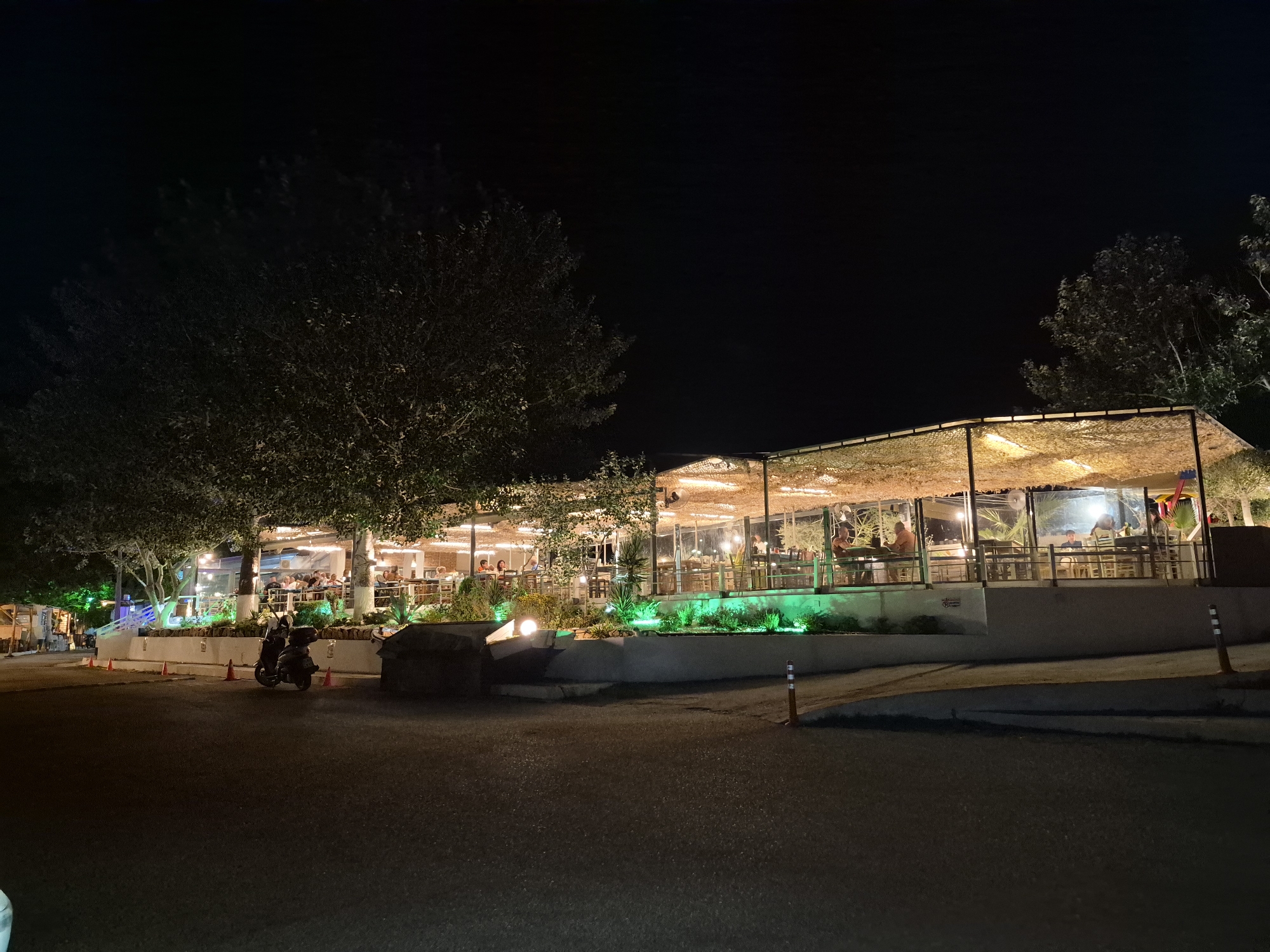 The image depicts a nighttime scene of a restaurant in Makri, Greece. The restaurant features an open-air seating area with a thatched roof, illuminated by warm lighting. Green lights highlight the landscaping in front of the building, creating a vibrant contrast with the dark sky. Several trees surround the restaurant, their foliage casting shadows. In the foreground, a paved area with traffic cones suggests a street or parking lot. Two scooters are parked on the paved surface. Inside the restaurant, patrons are seated at tables, engaged in dining and conversation. The overall atmosphere is one of a lively evening establishment.