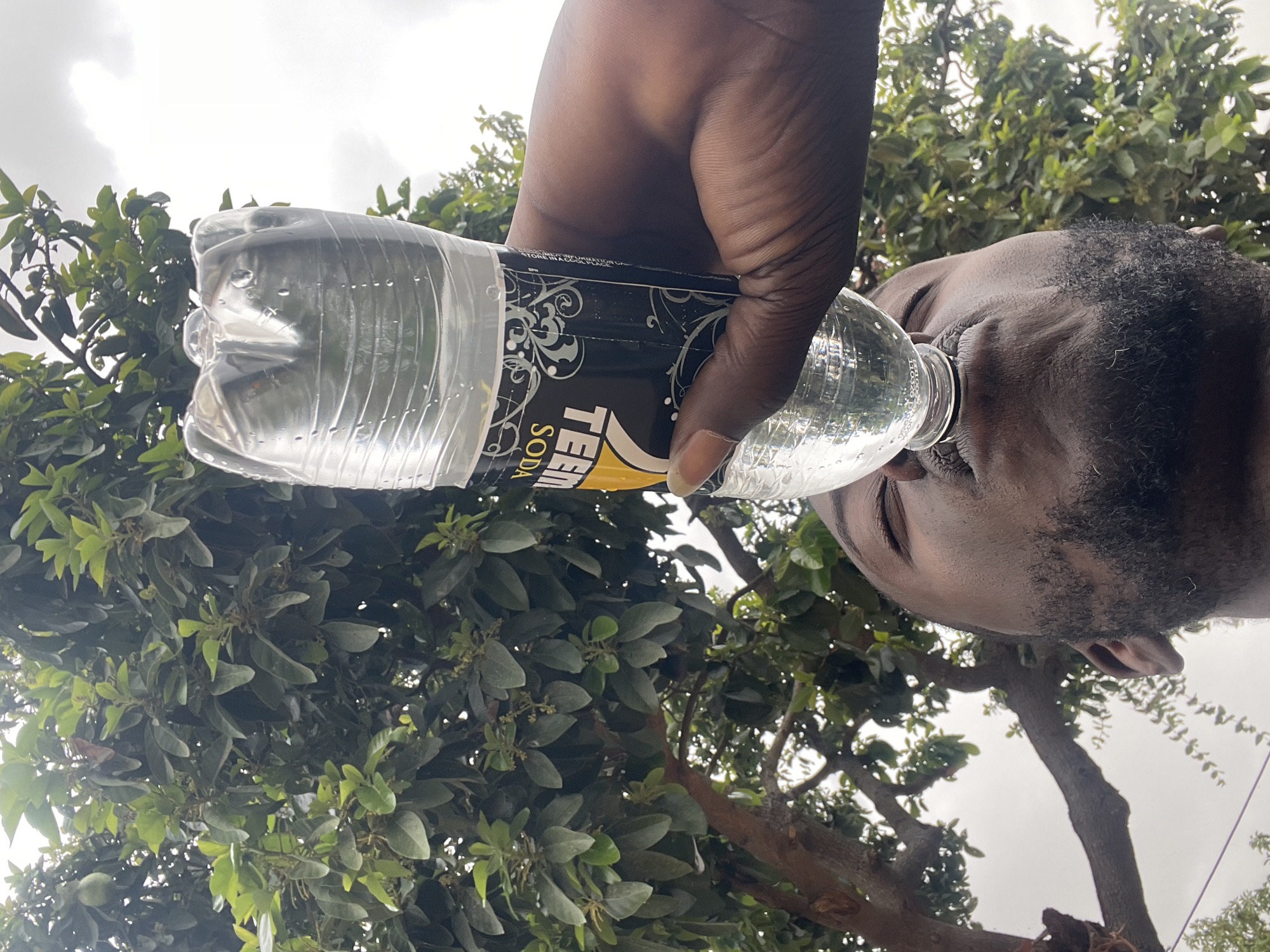 A man is drinking from a plastic soda bottle with the brand "TEEM SODA" visible on its label. The man's head is tilted back as he holds the bottle above him. His skin is dark and his hair is short and black. The bottle is clear and filled with liquid. The background consists of green foliage and trees, suggesting an outdoor location. The location is in the city of Unknown, Unknown.