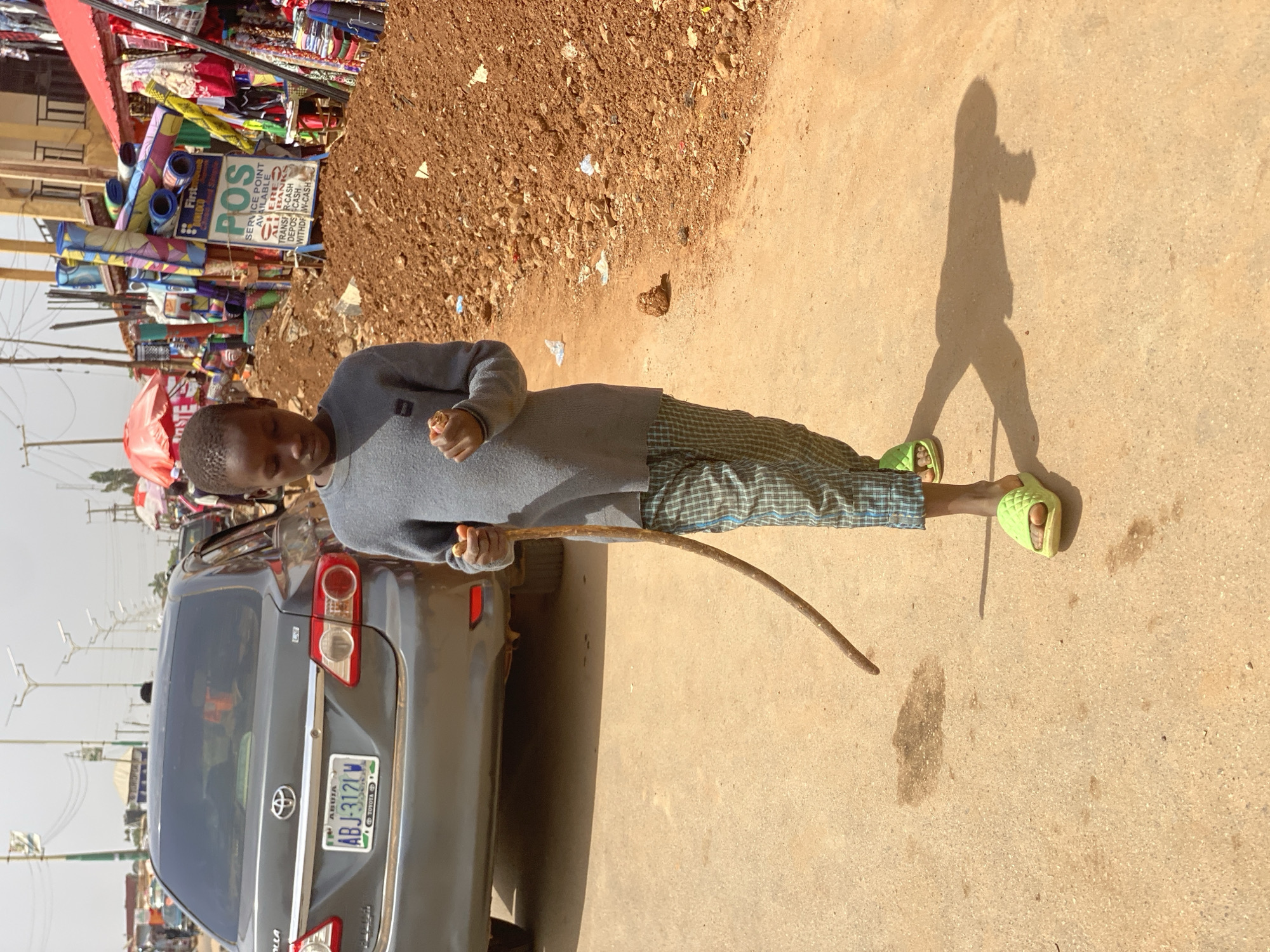 A young person stands on a paved surface next to a grey Toyota Corolla in Kafanchan B, Nigeria. The person is wearing a grey long-sleeved shirt, plaid trousers, and green sandals, and is holding a curved, slender stick. Behind the person is a pile of brown earth and a stall with fabrics and a sign indicating a "POS" (Point of Sale) service. The license plate of the car reads "ABJ-3121W" and indicates "ABUJA" on the Nigerian plate. The shadow of the person is cast on the paved surface. In the background, there are street lights and buildings along the road.