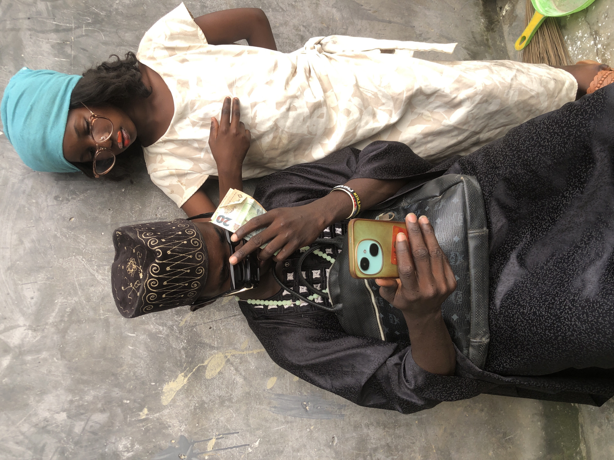 In Ado, Nigeria, a person wearing a black traditional outfit and a decorative cap is seated and looking at a smartphone in their hands. They are also holding a 20-unit banknote. Standing beside them is a child wearing a light-colored dress and a turquoise headscarf, with glasses on their face and orange lipstick applied. The child's hand is resting on the seated person's arm. The background is a rough, gray concrete wall. A black bag with a monogram pattern is positioned in front of the seated person. A broom and a green dustpan are visible on the ground in the lower right corner.