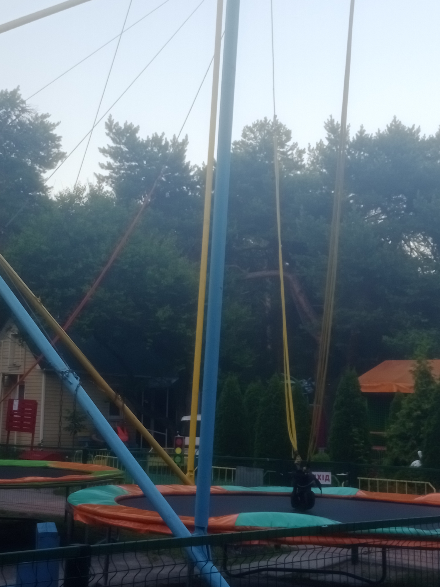 The image depicts an outdoor scene in Horishni Plavni, Ukraine, likely at an amusement or recreational park. Dominating the foreground are the structural elements of a bungee trampoline, including thick blue, yellow, and red elastic cords attached to upright poles, and the circular trampoline mats themselves, featuring orange and green surfaces. In the background, dense green trees form a natural backdrop, with some glimpses of man-made structures, including a light-colored building with a red sign and a building with an orange roof. The sky is overcast and bright. A figure is visible on one of the trampolines, appearing to be suspended by the bungee cords. There is a traffic light visible to the right. The overall impression is that of an amusement area set within a natural, wooded environment.