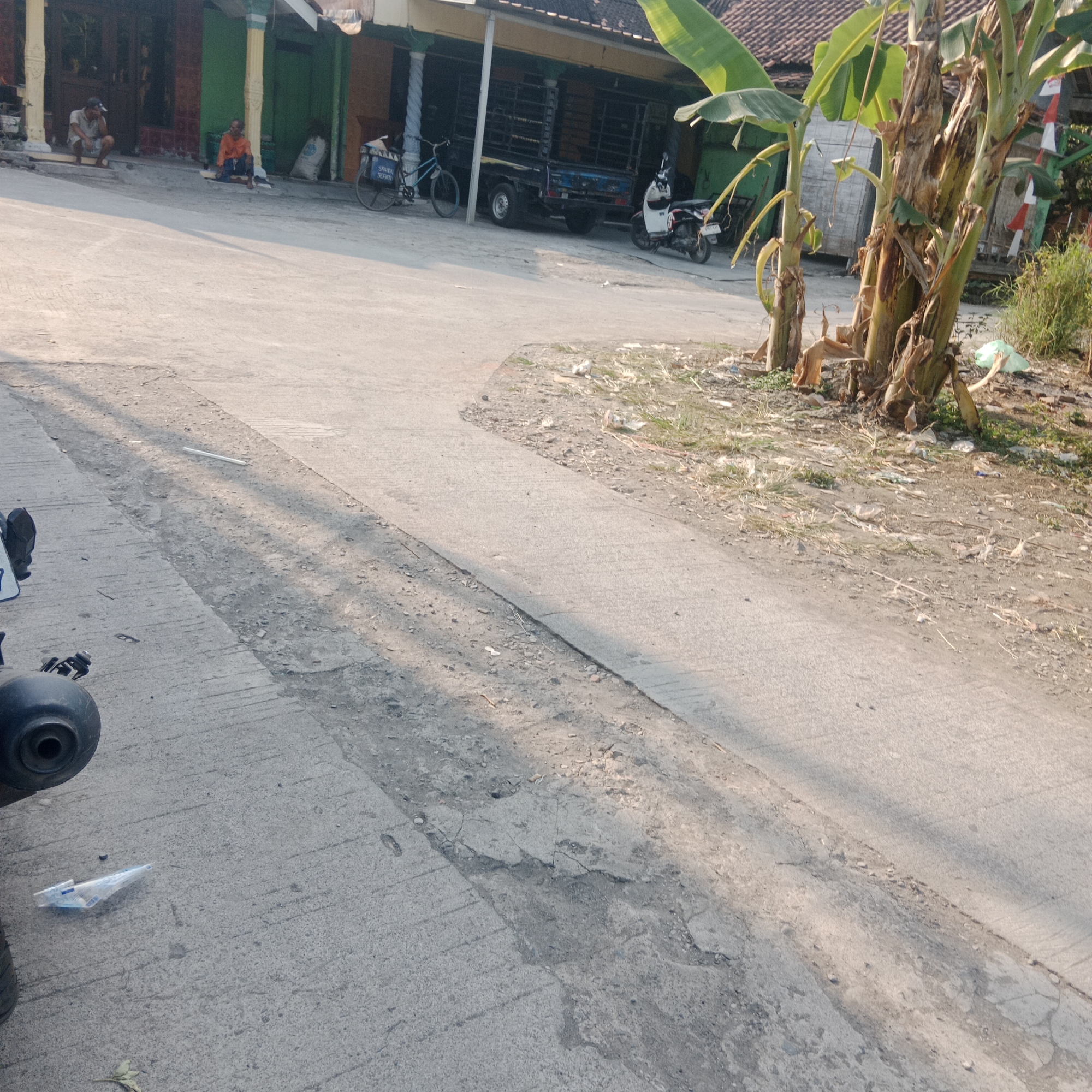 The image depicts a street scene in Tegowanu Kulon, Indonesia. In the foreground, the corner of a black motorcycle exhaust is visible on the left. A plastic bag lies on the concrete road. The road itself is made of concrete and shows signs of wear and tear, with some cracks and uneven patches. In the mid-ground, a white plastic item, possibly a water bottle or packaging, is present on the road. Further back, on the right side of the road, a clump of banana trees is situated near a patch of dry grass and some scattered debris. The background shows a building with green and yellow facades, a black truck parked in front, and a bicycle. Two individuals are seated on the steps in front of the building; one is wearing an orange shirt and the other a light-colored shirt and a cap. The lighting suggests it is either early morning or late afternoon, casting long shadows across the road.