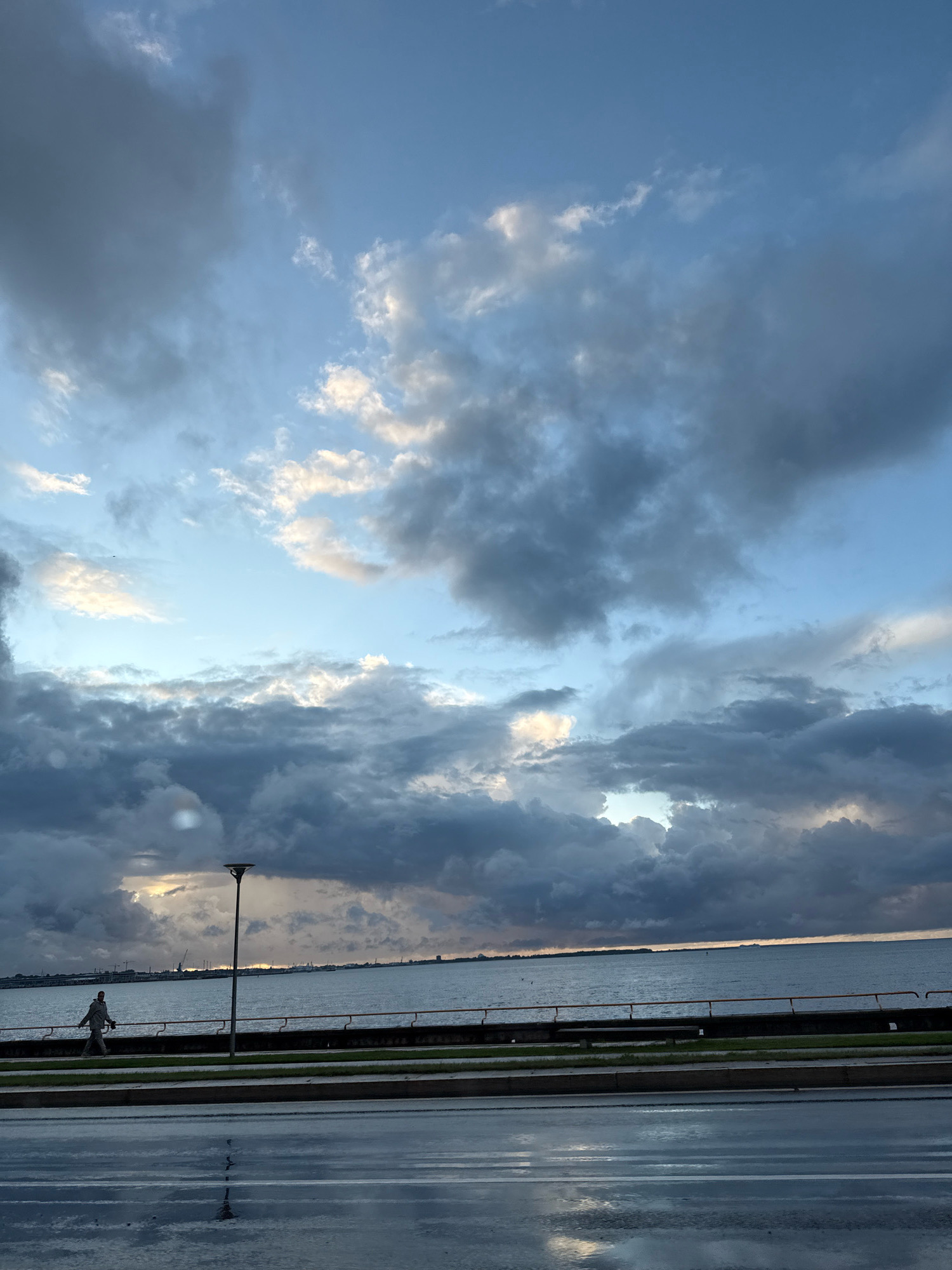 The image depicts a waterfront scene in Tallinn, Estonia, under a dramatic sky filled with dark, heavy clouds interspersed with patches of lighter sky and illuminated clouds. In the foreground, a wet, reflective asphalt road stretches across the width of the frame, suggesting recent rain. Beyond the road, a strip of grass separates it from a paved promenade. A solitary figure, appearing to be a person walking, is visible on the promenade, moving from left to right. A tall lamppost stands near the promenade's edge, its dark silhouette contrasting with the sky. The promenade runs parallel to a body of water, likely the Baltic Sea, which is calm and stretches towards the horizon. On the distant horizon, the faint outline of buildings and industrial structures can be observed, characteristic of a port city. The overall atmosphere is moody and overcast, with the play of light and shadow on the clouds creating a sense of depth and impending weather change.