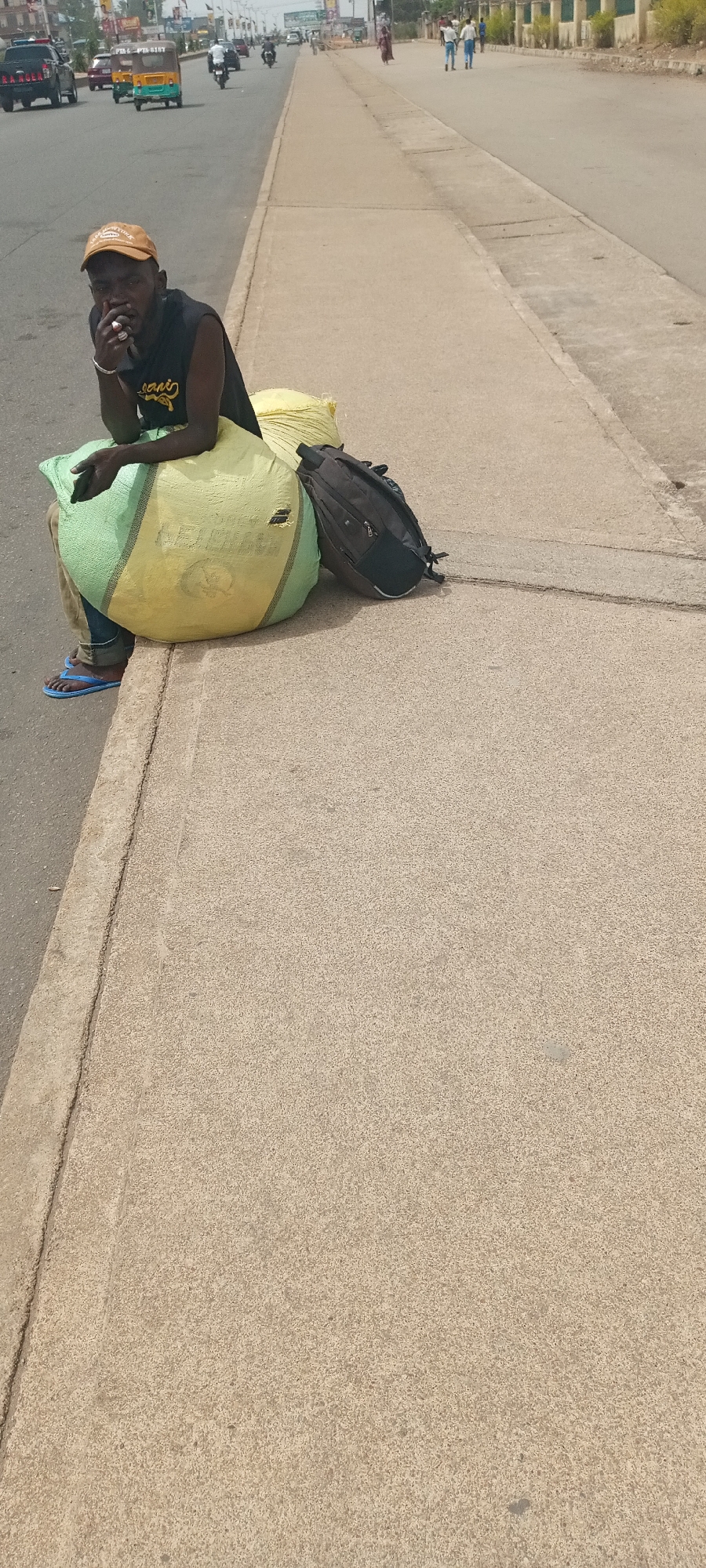 A man with dark skin, wearing a yellow cap, black tank top, and blue sandals, is sitting on a large, green and yellow sack on a concrete curb next to a road. He appears to be smoking, holding something to his mouth with his right hand. A yellow sack and a black backpack are next to him. The background shows traffic and pedestrians on a busy road in Jos, Nigeria.