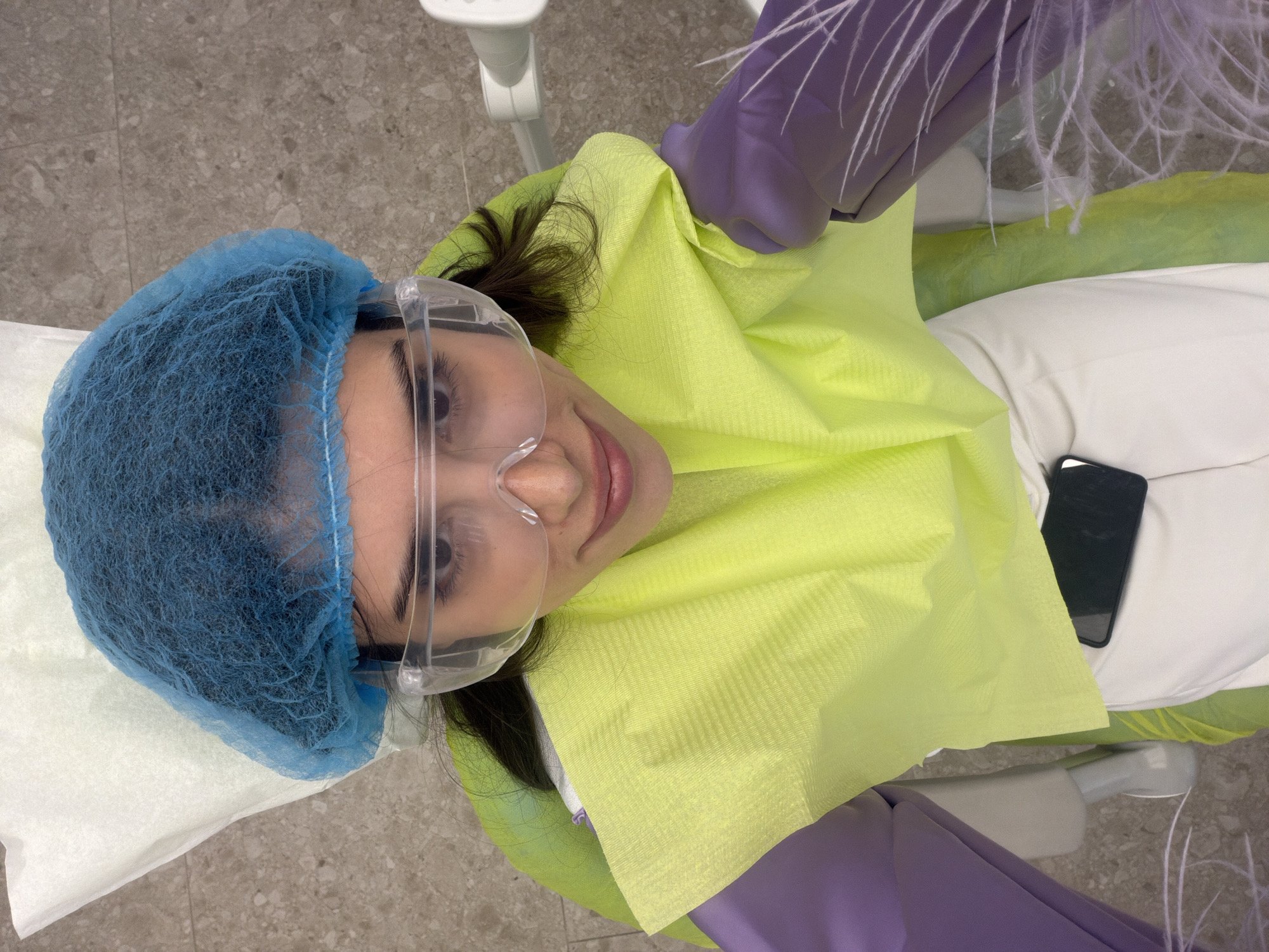 A person is lying down on a dental chair, wearing a blue surgical cap, clear safety goggles, purple gloves, and a bright yellow bib. The person is looking up at the camera and smiling. A black smartphone is placed on their lap. The background shows a tiled floor and dental equipment. The scene appears to be a dental office.