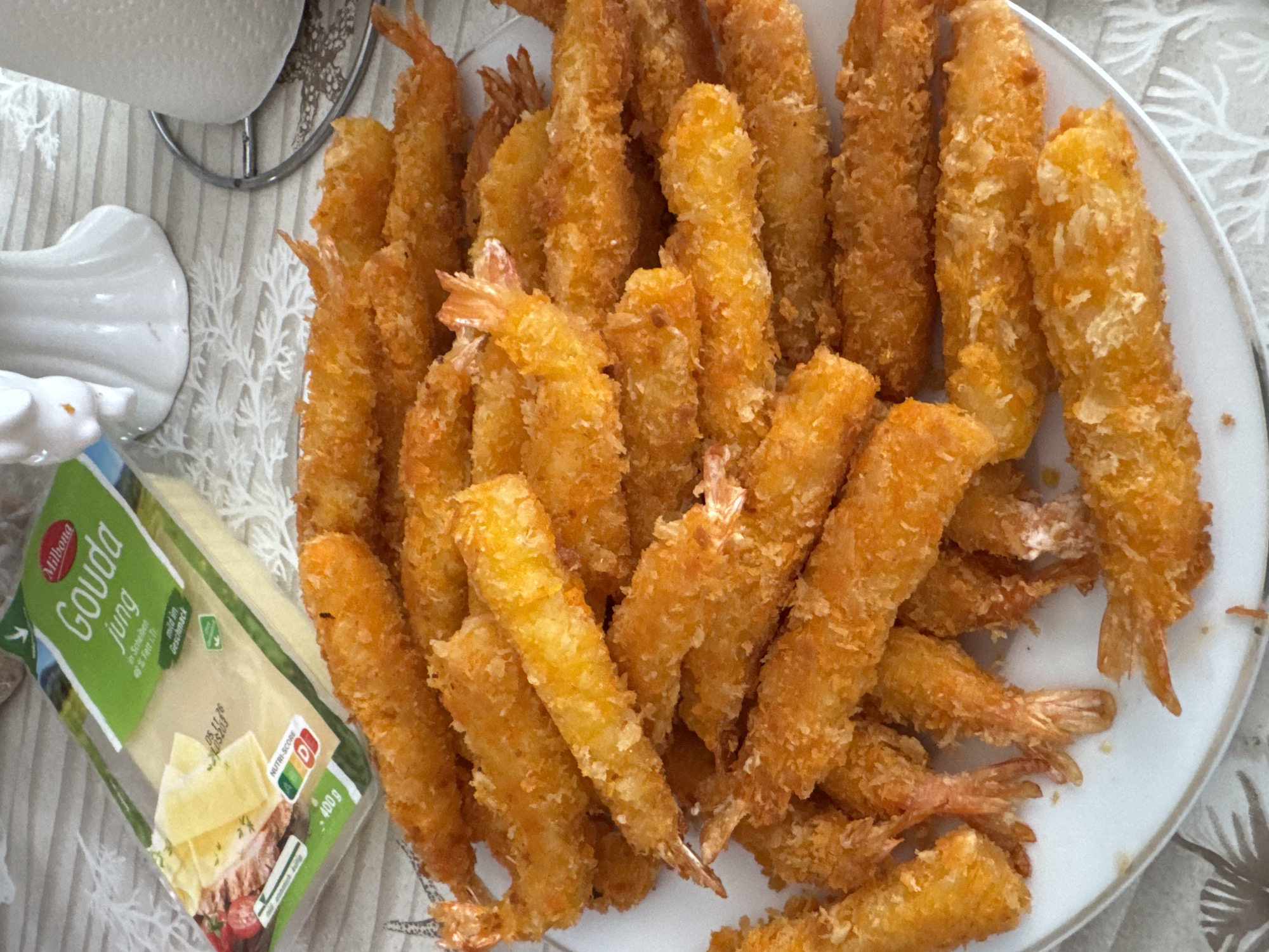 A plate of deep-fried shrimp, coated in breadcrumbs, is arranged on a white plate. The shrimp are golden brown and appear to be fresh. In the background, a package of Milbona Gouda cheese is visible. The overall scene suggests a meal is being prepared or served. The image does not provide information about the location in the city of Rheine, Germany, or any people present.