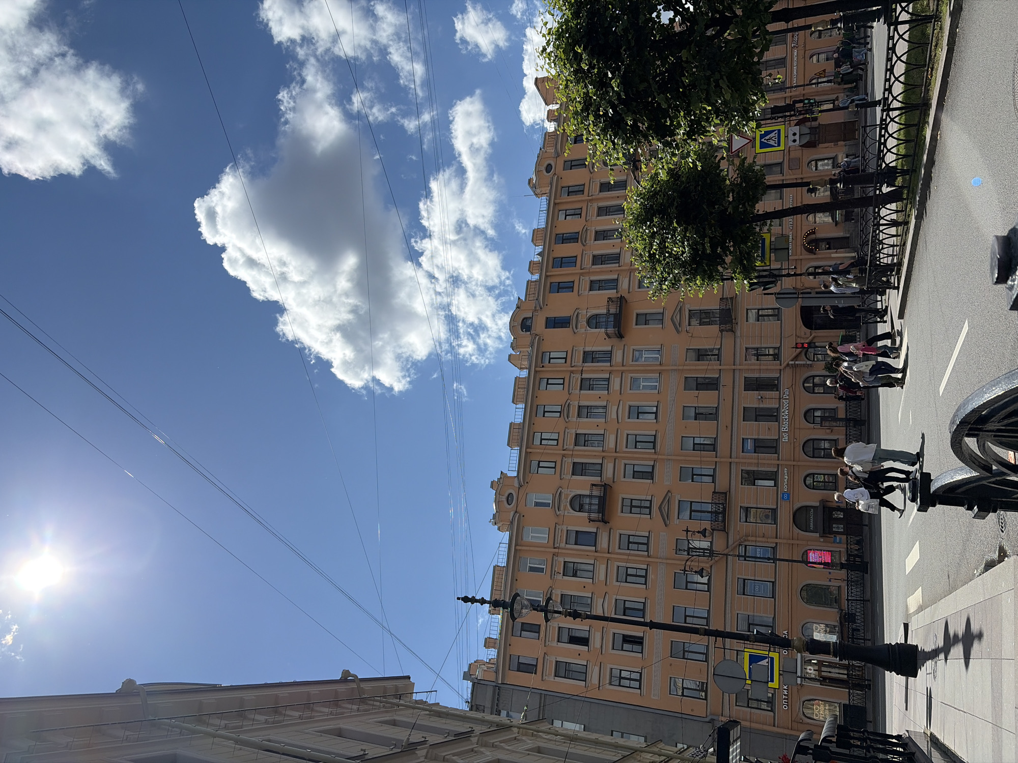 The image captures a sunny daytime scene on a street in Saint Petersburg, Russia, likely near Nevsky Prospect, a prominent avenue. The main subject is a large, multi-story building with a peach-colored facade, featuring numerous windows with dark frames. In front of the building, a street with crosswalks is visible, with several pedestrians crossing.
The sky is bright blue with a few fluffy white clouds and a strong sun, creating lens flare in the upper left quadrant. Overhead, a network of utility wires crisscrosses the sky. Lampposts, one of which is ornate and black, line the sidewalk. Trees with lush green foliage are planted along the street, some obscuring parts of the building.
On the building's ground floor, businesses are indicated by signage. One sign reads "ОПТИКА" (Optics), another appears to say "КОПИЦЕНТР" (Copy Center), and further down, "BlackWood Pub" is visible. Another sign, partially obscured by a tree, seems to advertise "МАГАЗИН ДЕТСКОЙ КОСМЕТИКИ" (Children's Cosmetics Store). Yellow pedestrian crossing signs are also present at the intersections.
People are seen walking across the street, dressed in casual clothing, suggesting a typical urban environment. The architecture of the building, with its ornate details and repeating window patterns, is characteristic of Saint Petersburg's historical city center.