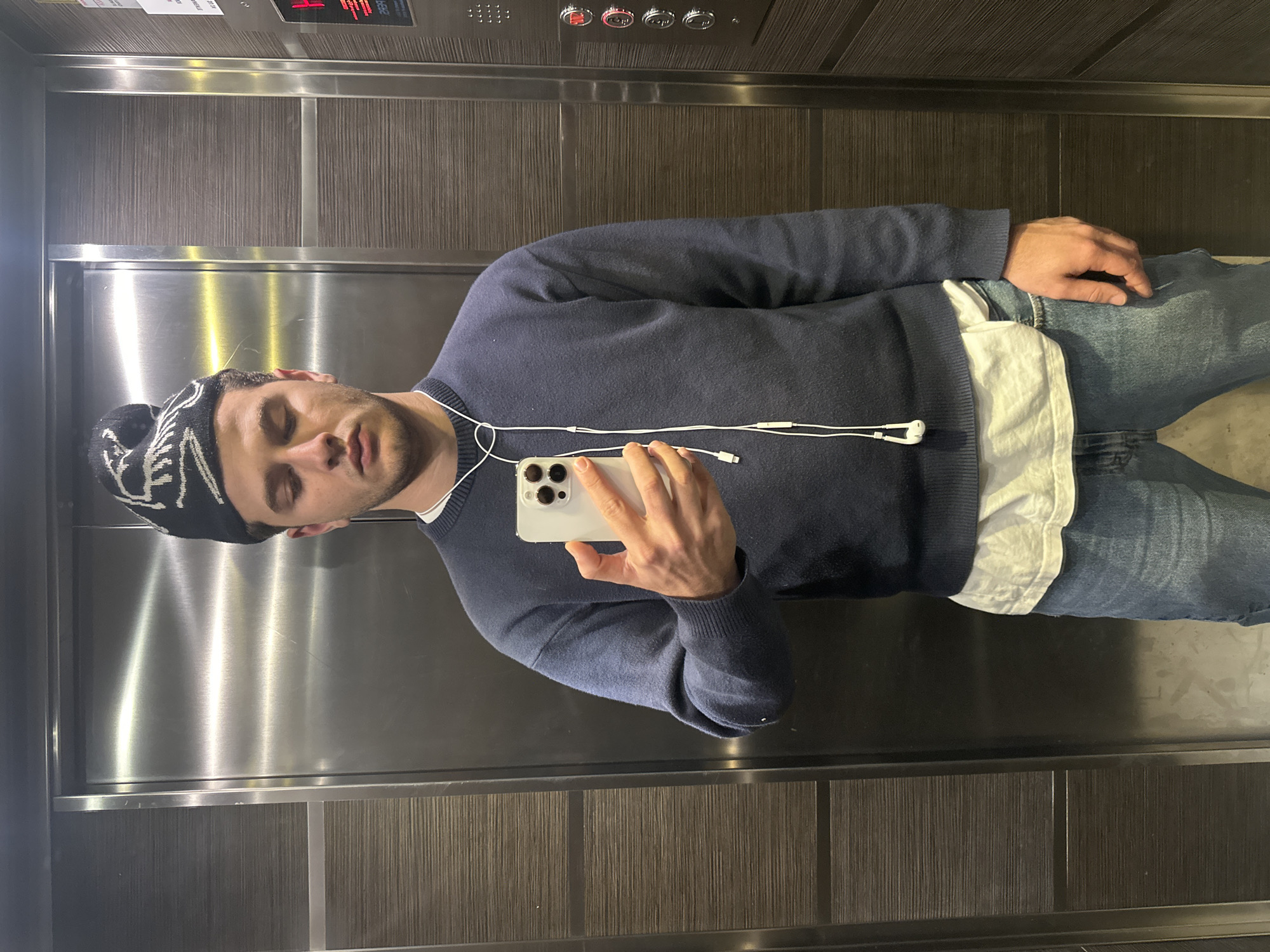 A man is taking a selfie in an elevator. He is wearing a dark blue sweater, light blue jeans, a white t-shirt peeking out from under the sweater, and a black and white patterned beanie. He is holding a white smartphone and has white wired earphones plugged in. The elevator has a brushed metal interior with a dark wood-paneled section. The location is unknown, and there is no indication that the image was taken in Milan, Italy.