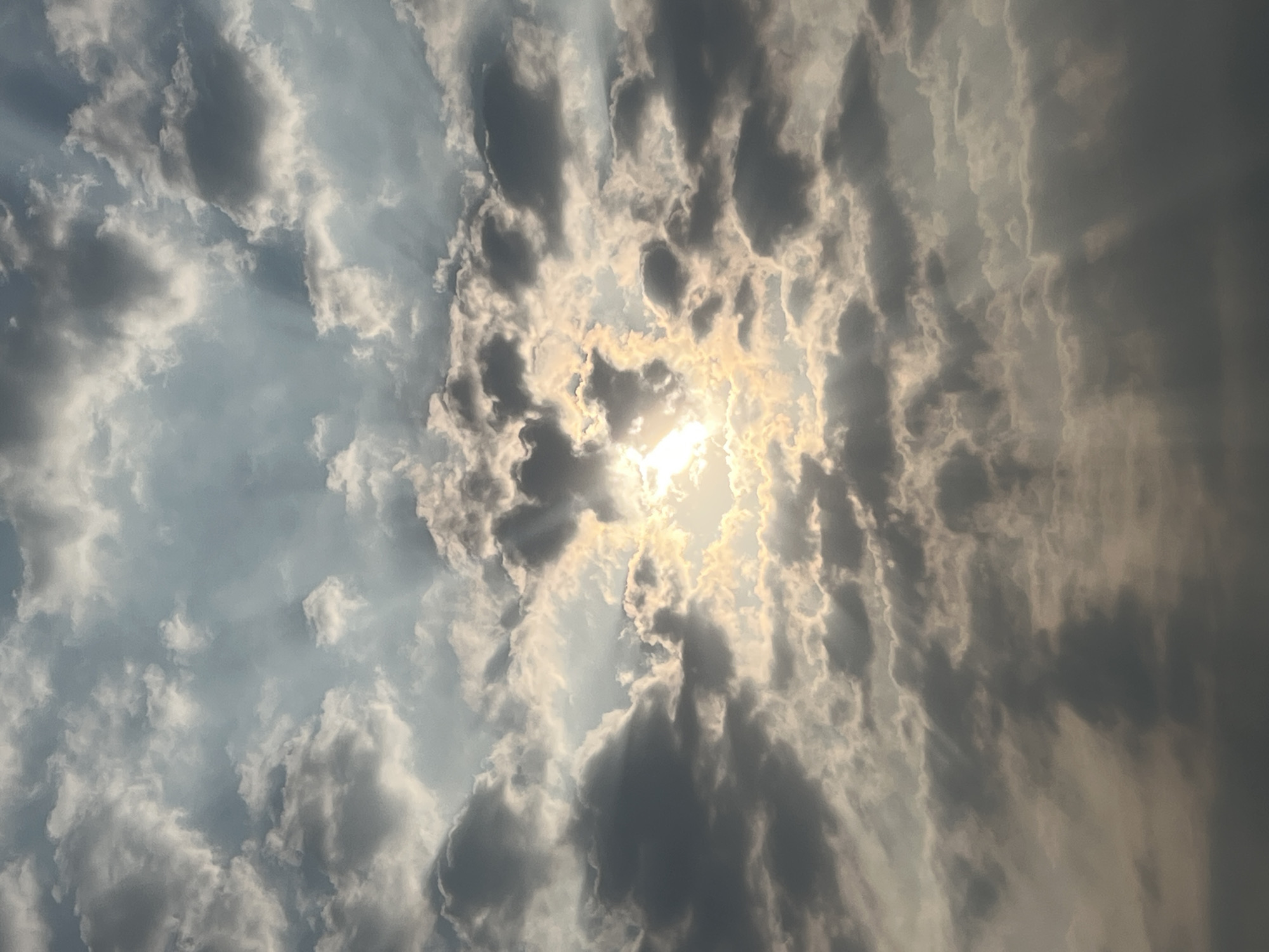 The image depicts a sky view with the sun partially obscured by clouds. The scene displays a mix of light and dark clouds with sun rays piercing through the gaps. The location details are not visually discernible from the provided image. There are no visible people, subjects, or objects apart from the sky and clouds. No specific actions or interactions are observed. The environment is limited to the sky and atmospheric elements captured in the image.