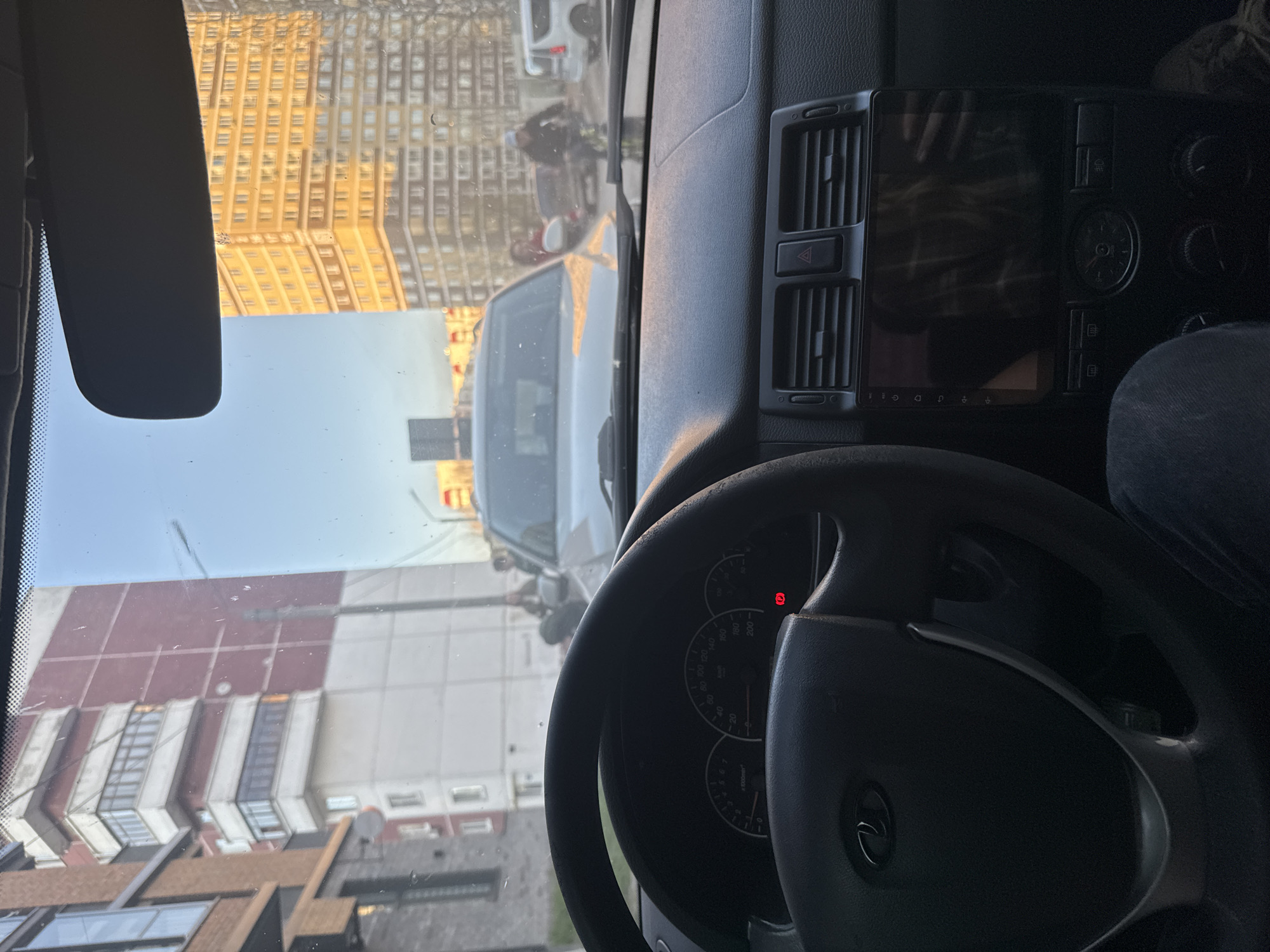 The image shows the interior of a Lada automobile, with a view through the windshield of buildings in what appears to be an urban setting. Specifically, the location is Perm, Russia. In the foreground, the car's dashboard and steering wheel are visible. The dashboard features a radio/infotainment system with a digital display, hazard lights button, and climate control dials. The steering wheel has the Lada logo at its center, and the instrument cluster shows a speedometer with markings in kilometers per hour.
Outside the windshield, there is a street with other vehicles and pedestrians. Buildings of varying colors and architectural styles line the street. One building has a mix of reddish-brown and white facade.
