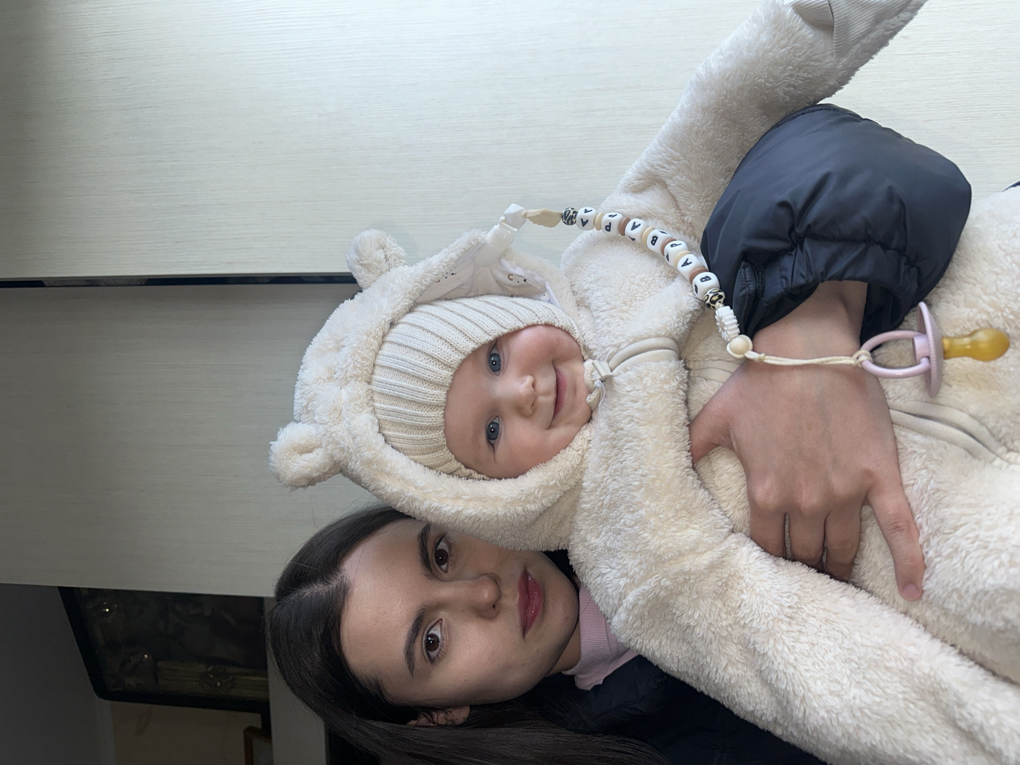 A medium shot captures a woman holding a baby. The baby wears a cream-colored fuzzy bear suit with a matching knit hat and has a pacifier attached to the suit with a beaded chain. The woman has long, dark hair and dark eyes. They are positioned indoors, likely in a home, with a white and light gray table surface visible in the background. The location is assumed to be Chaikovsky, Russia.