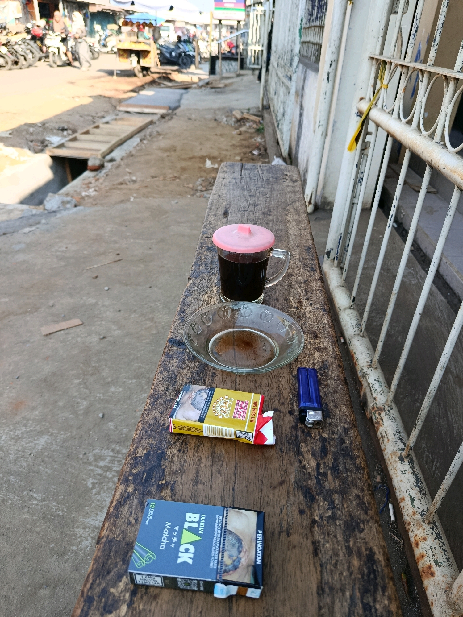 The image depicts an outdoor scene on a wooden bench, likely in a semi-urban or rural area, possibly a roadside stall or market in Bululawang, Indonesia. A clear glass mug filled with a dark liquid, likely coffee, sits on the bench, topped with a pink lid. Next to it is a small, clear glass saucer with residue. In front of the mug are two open packs of cigarettes, one with yellow and red packaging featuring text in Indonesian, and another with black and green packaging also displaying Indonesian text and nicotine content information. A blue disposable lighter lies between the cigarette packs. The background reveals a dusty street with motorcycles parked, a cart, and construction or repair work in progress, indicated by a partially constructed wooden structure over an open drain. The overall environment suggests a casual setting, possibly during daylight hours due to the bright lighting.