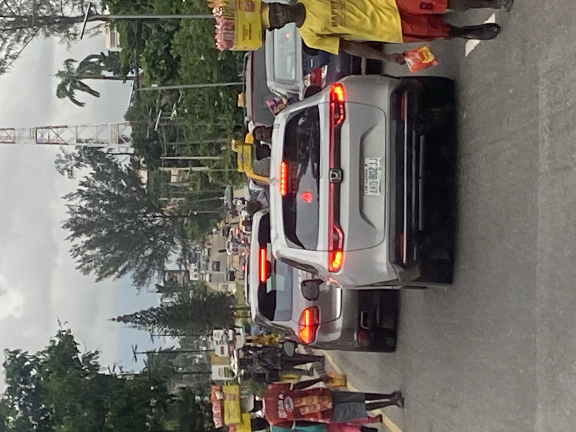 In Ikeja, Nigeria, a traffic jam unfolds on a cloudy day. Several cars are stationary on the asphalt road, with brake lights illuminated. Pedestrians navigate the congested street, some carrying goods on their heads. A man in a red shirt and grey shorts walks with a yellow package in his hand, while another man in a yellow t-shirt and red shorts holds a red package. Trees line the sides of the road, and a telecommunications tower is visible in the background. Traffic lights and street lamps are also present.