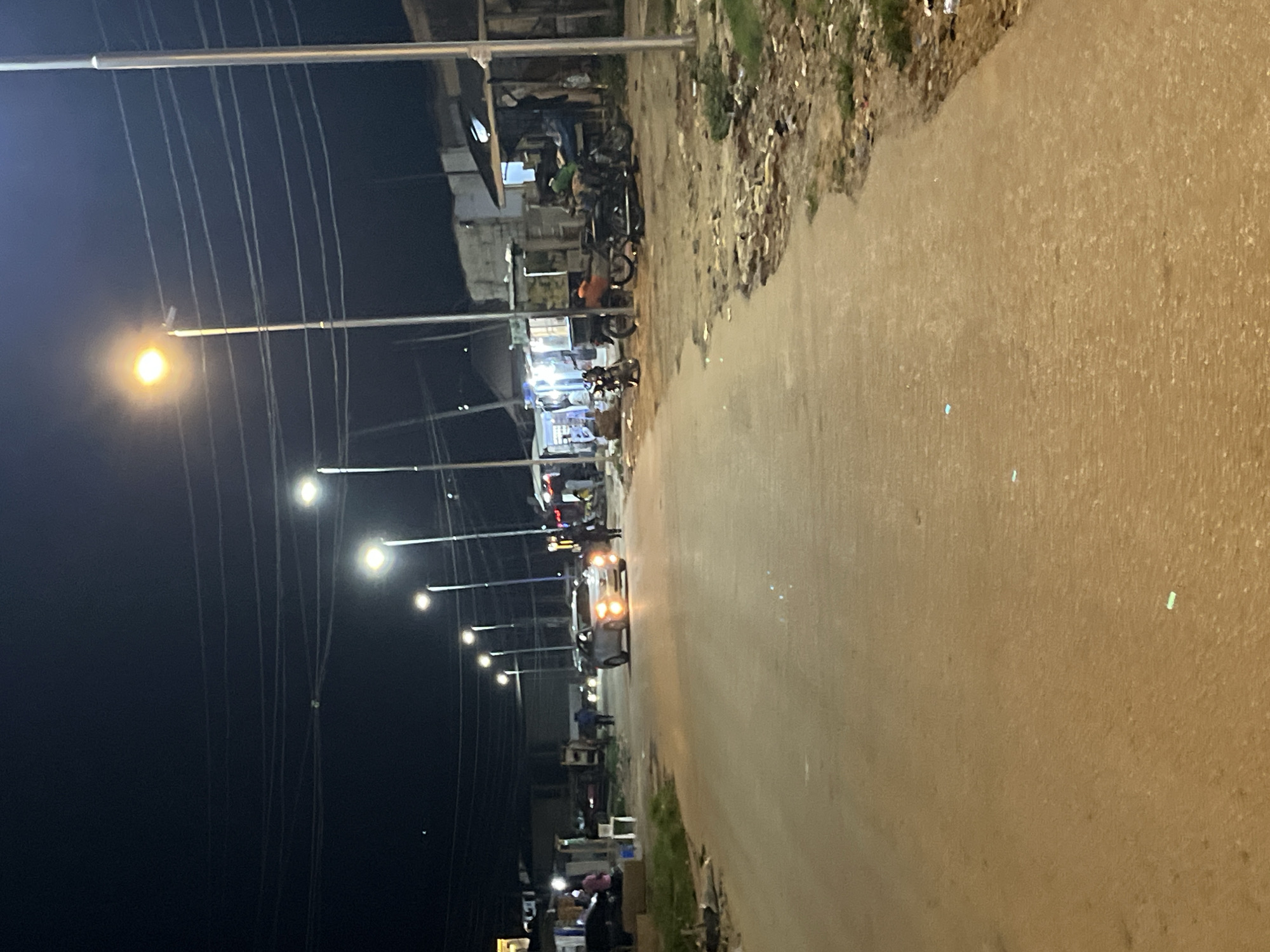 The image depicts a street scene at night in Abuja, Nigeria, illuminated by streetlights. A white SUV is driving down the road, with its headlights on. Several motorcycles are parked on the side of the road, and there are buildings and shops lining the street. The road appears to be paved, and there is some debris and vegetation along the sides. The overall impression is of a typical urban street at night, with signs of commercial activity.