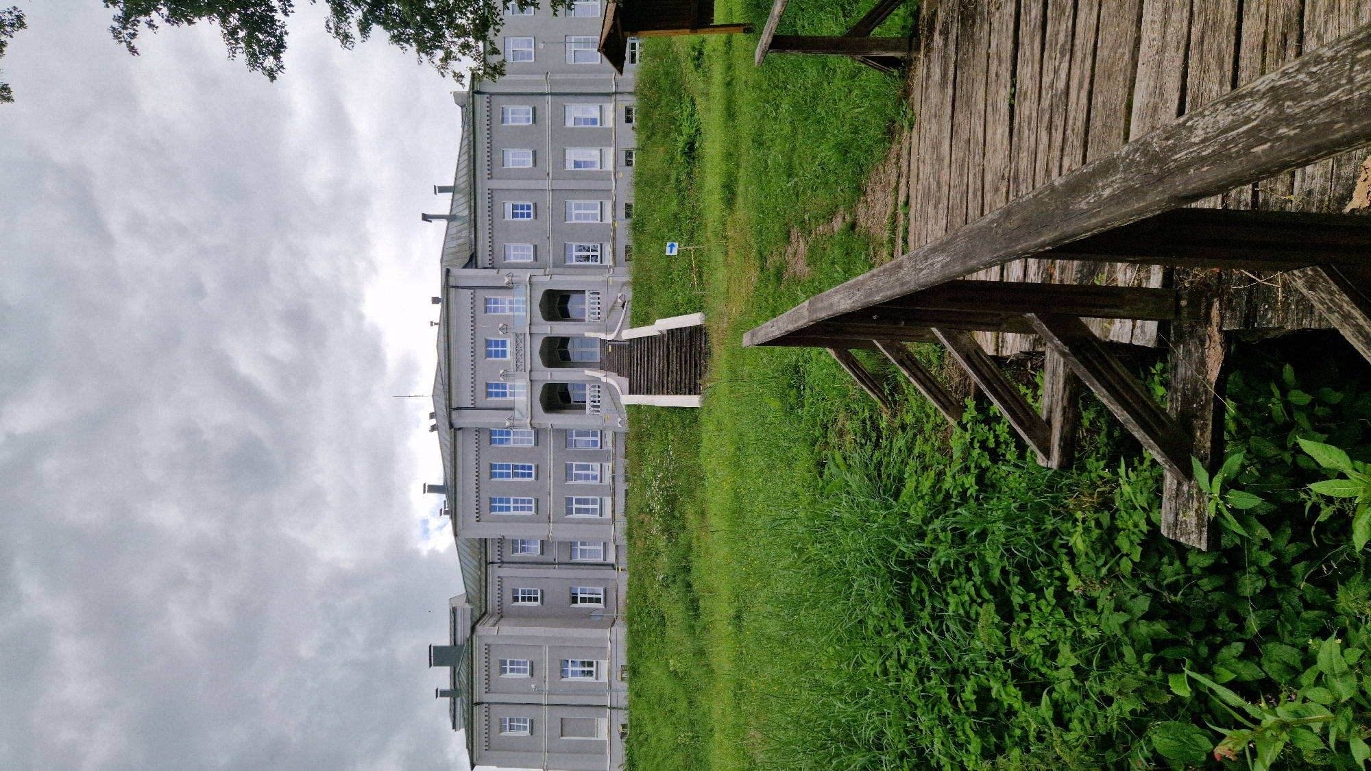 The image shows a wide, grey, two-story building with a symmetrical facade and a central entrance featuring a covered balcony. The building is surrounded by a lush green lawn that slopes upwards towards it. A set of stone stairs leads from the lawn to the building's entrance. In the foreground, a wooden bridge with a weathered texture and railing extends into the frame. The sky is overcast with grey clouds. A blue directional arrow sign is visible on the lawn, pointing towards the building. The scene appears to be outdoors in a park-like setting. This building is located in Priekule, Latvia.