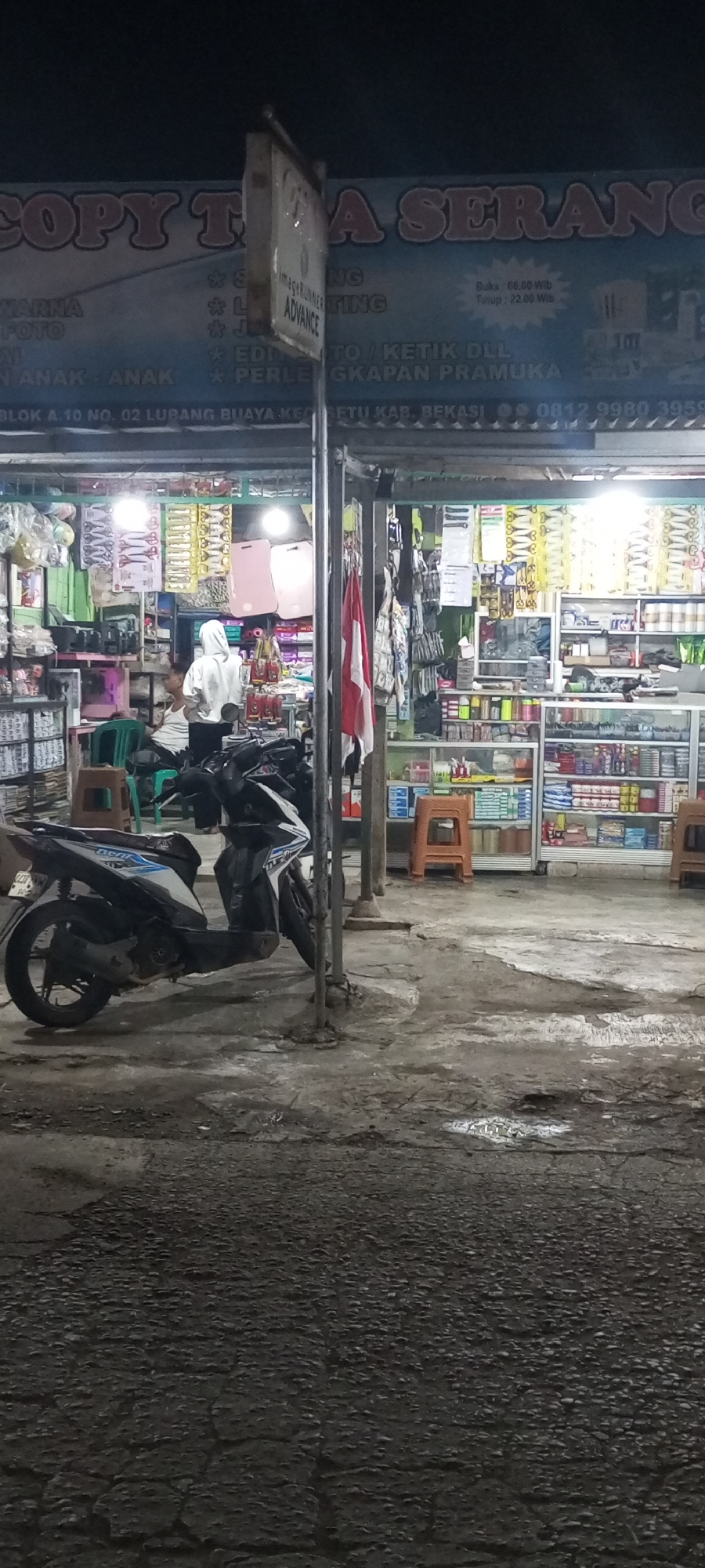 This is a photograph of a storefront in Setu, Indonesia, taken at night. The storefront appears to be a copy and print shop that also sells stationery and other supplies. A large sign above the entrance reads "COPY TUA SERANG" and lists services such as "WARNA FOTO," "JILID," "SCAN," "EDIT FOTO," "KETIK DLL," and "PERLENGKAPAN PRAMUKA." The shop is open as indicated by the sign displaying "Buka: 06.00 Wib" and "Tutup: 22.00 Wib."
Inside the shop, shelves are stocked with various items, including what appear to be paper, notebooks, pens, and craft supplies. Two individuals are visible inside; one is wearing a white hooded sweatshirt and appears to be standing near the counter, while another individual is seated to the left.
In front of the shop, two motorcycles are parked on a wet, uneven asphalt surface. The area is dimly lit by the shop's interior lights and possibly some streetlights. The overall impression is of a functional, everyday business operating in a residential or mixed-use area.