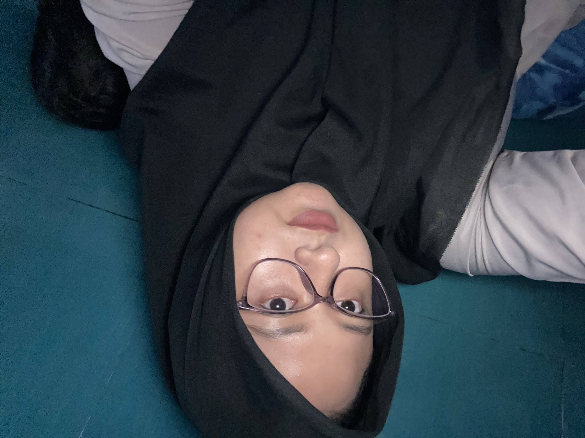 The scene is located in Gelam, Indonesia. The image features a young adult female positioned in a self-portrait, looking directly forward into the camera. She wears a black hijab that frames her face and a light-colored, likely white or off-white, long-sleeved garment visible at the shoulders and arms. Her face has fair skin, and she wears eyeglasses with light purple frames. Her lips are adorned with a light pink or nude lipstick. The background consists of a solid teal-colored wall, exhibiting minor textural variations and faint streaks. To the lower left, a blue fabric with an indistinct pattern is partially visible. On the lower right, behind her right shoulder and partially obscured by her clothing, rests a dark, textured object. The lighting appears direct and frontal, creating reflections in the eyeglasses and highlights on the forehead and nose.