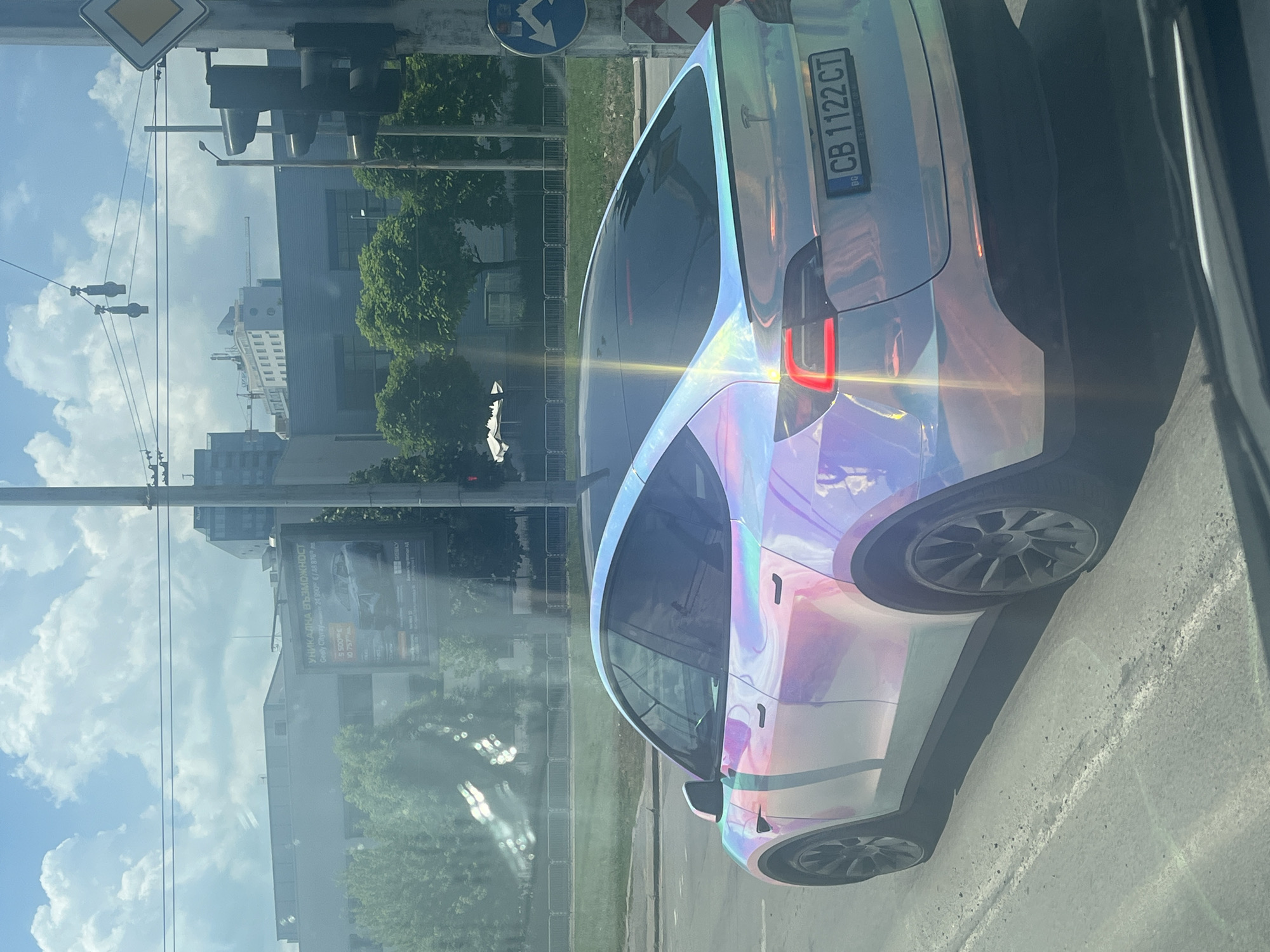 The image shows a Tesla Model Y with a holographic wrap parked on a city street. The car is white with iridescent pink, blue, and green colors reflecting off its surface. The license plate reads "CB 1122 CT". In the background, there are buildings, trees, and a traffic light. The sky is cloudy. The image appears to have been taken from another vehicle, as evidenced by the reflection of the dashboard in the foreground.