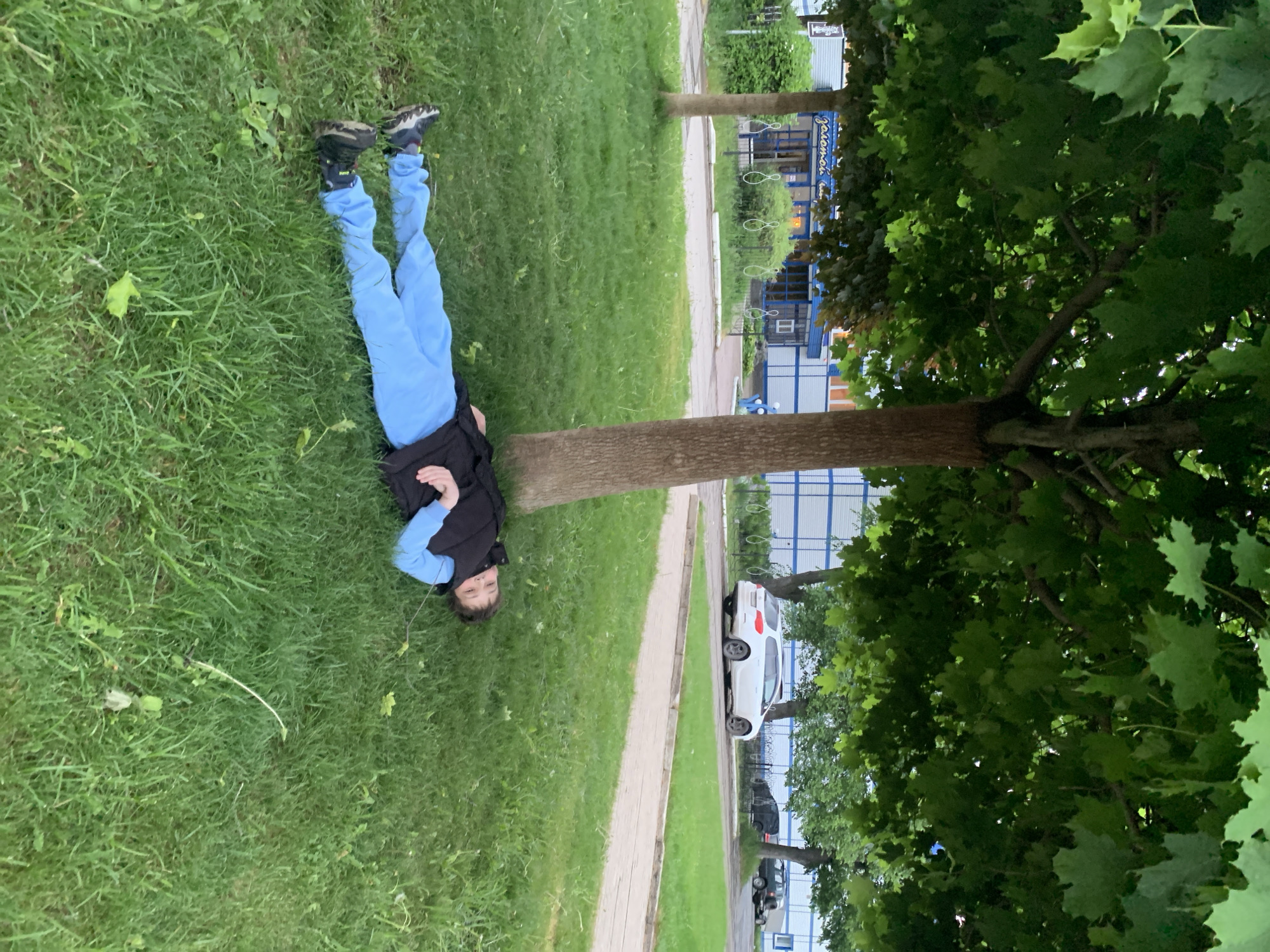 A child is lying on their back in a grassy area, possibly a park, in Chaikovsky, Russia. The child is wearing blue pants, a light blue long-sleeved shirt, and a black vest. Their feet are pointing towards the top left of the frame. A tree trunk is partially in the middle of the image, with its leaves forming a canopy on the right side. Behind the tree, across a walkway, are buildings, including what appears to be a storefront or public building with blue and white striped paneling. There are parked cars further back and down the road.