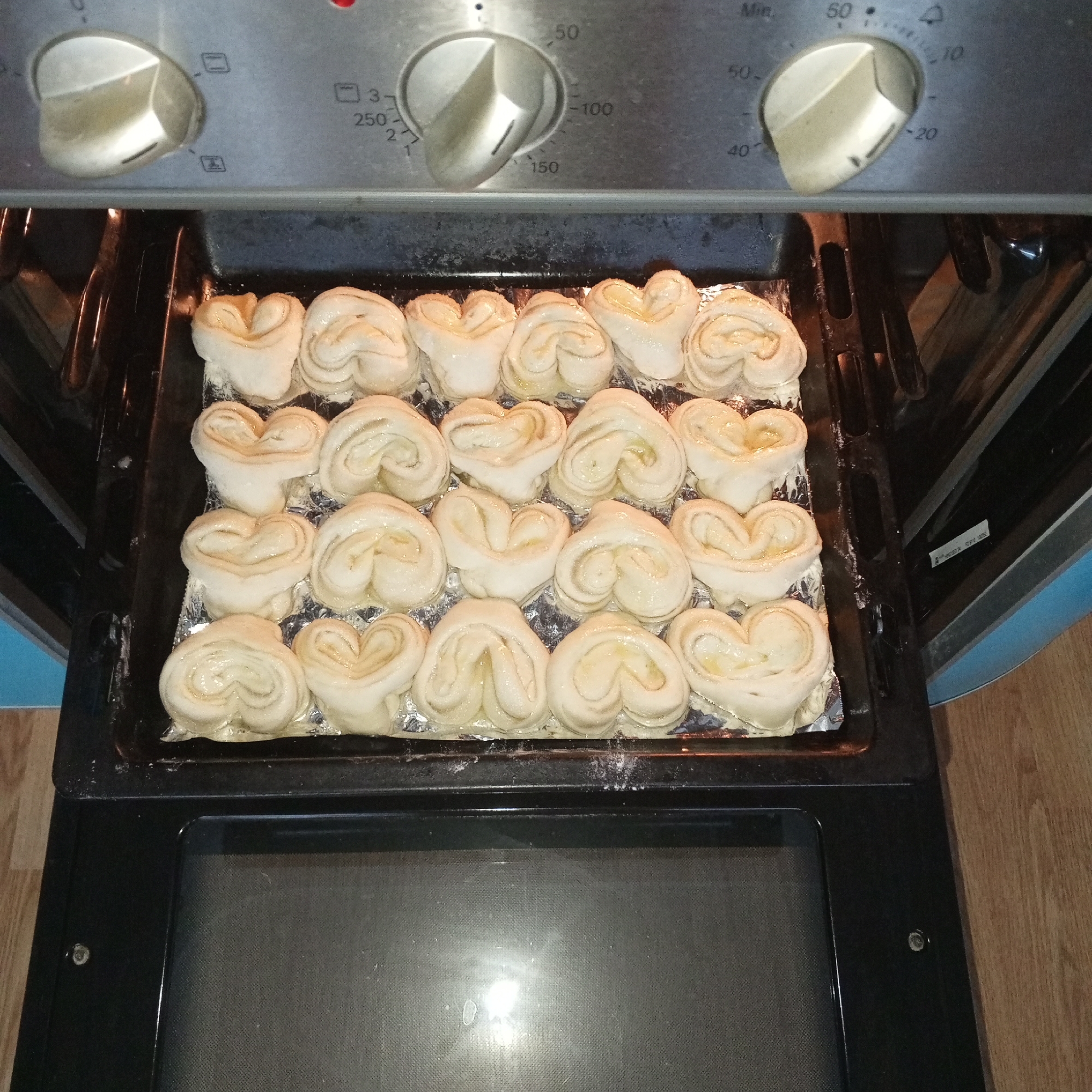A tray of raw, heart-shaped pastries rests on a foil-lined baking sheet inside an oven. The oven's knobs are visible, showing settings for temperature and timer. The oven is located indoors. There is no way to determine if the oven is in Gremyachinsk, Russia.