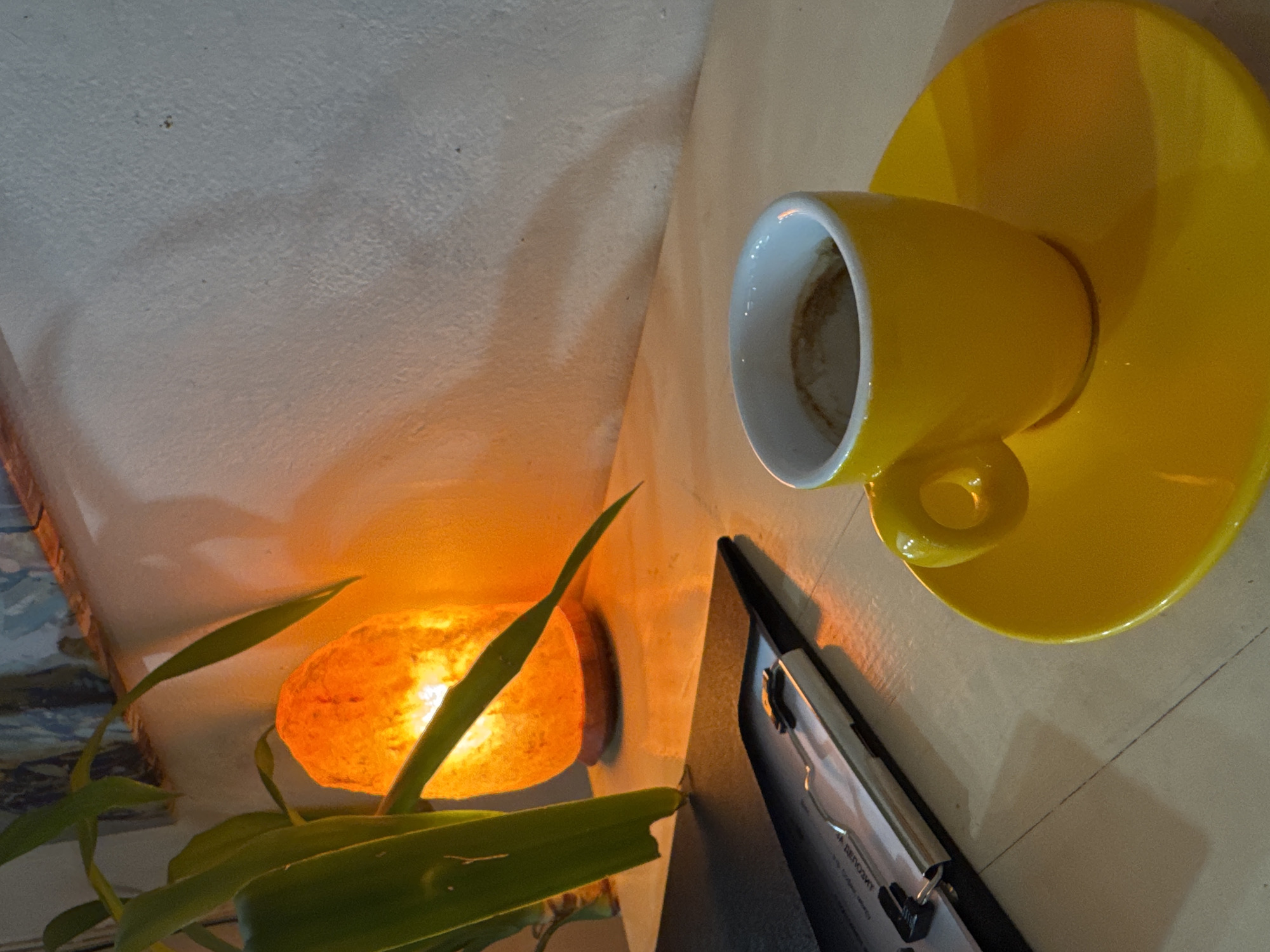 This image presents an indoor still life, set in Sofia, Bulgaria, as indicated by visible Bulgarian text. The scene features a bright yellow coffee cup resting on a matching saucer, positioned towards the upper right. The cup's white interior shows traces of a consumed beverage.
To the left, a glowing orange Himalayan salt lamp illuminates a textured, light-colored wall, casting a warm ambiance. A green potted plant with long, slender leaves partially obscures the salt lamp, its foliage reaching into the foreground and silhouetted against the light.
Below the coffee cup and to the right of the plant, a dark clipboard with papers is visible. Partially legible text on the clipboard clearly shows the phrase "HA DЕПОЗИТ," which is Bulgarian for "ON DEPOSIT," serving as a definitive location cue for Sofia, Bulgaria. The objects are arranged on a light-colored surface, possibly a table or countertop. The overall lighting, primarily from the salt lamp, suggests the photograph was taken in a dimly lit environment, likely evening or night, contributing to a calm and cozy atmosphere. No individuals are present in the image.