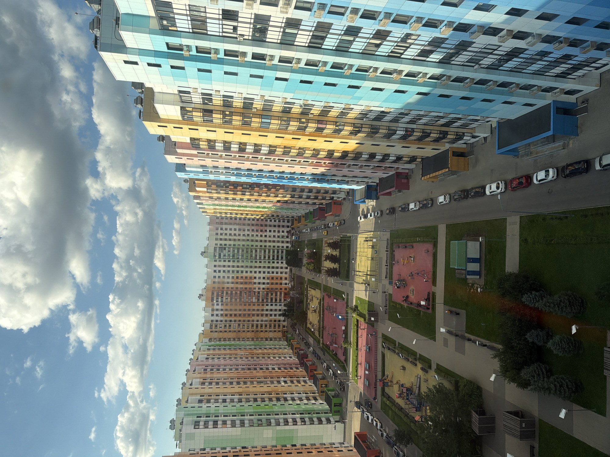 This is a high-angle, eye-level shot of a brightly colored, modern residential complex in an urban environment. The buildings are tall and multi-story, with facades painted in a variety of vibrant colors, including pastel shades of green, orange, pink, yellow, and blue, arranged in a repeating pattern. The buildings are lined up along streets, creating a sense of depth and perspective. In the foreground and middle ground, there are well-maintained courtyards with playgrounds, sports facilities, and green spaces, including trees and grass. Several parked cars are visible along the streets and in designated parking areas. The sky is a clear blue with scattered white clouds, suggesting daytime with good weather. The overall scene depicts a well-planned and visually appealing urban living space.