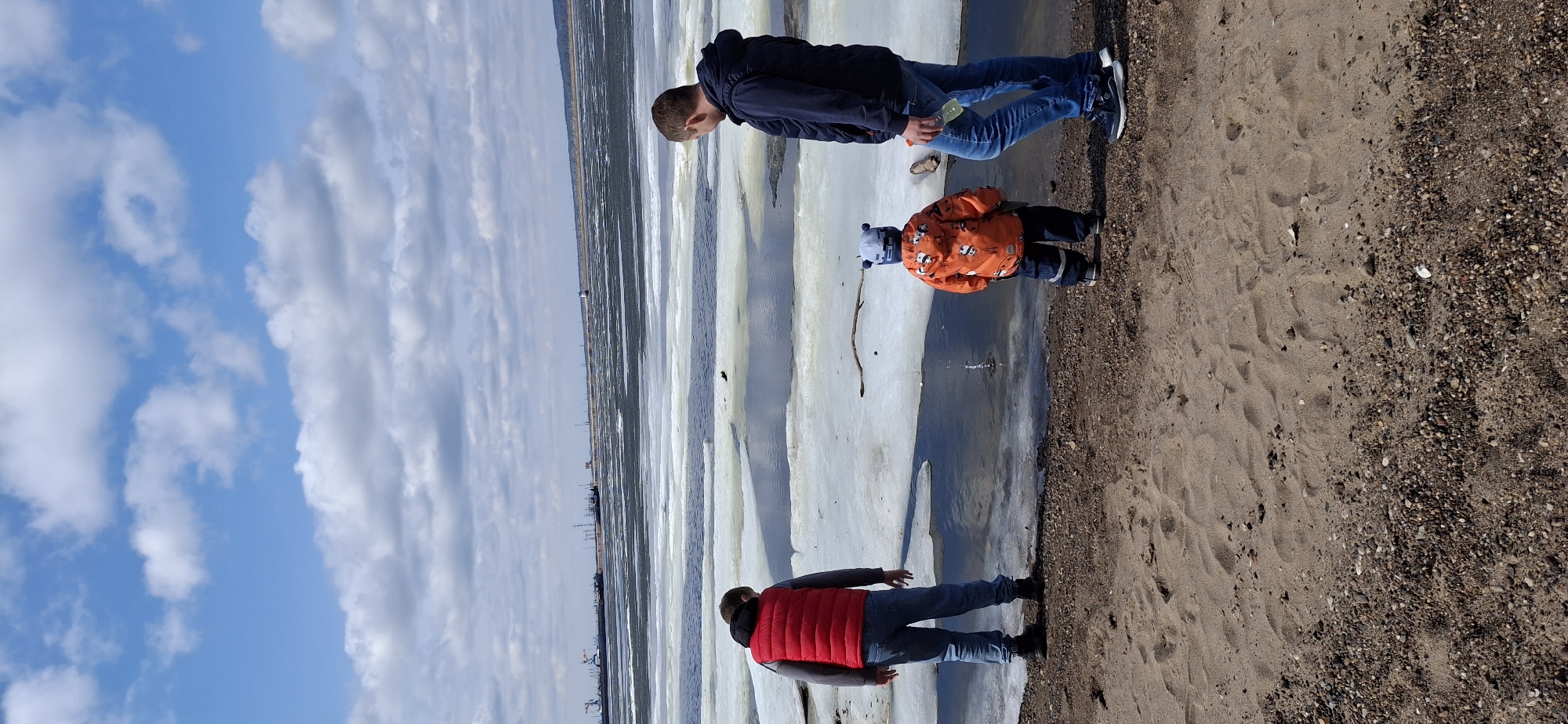 Three individuals stand on a beach in Chaikovsky, Russia. The beach has a sandy section that transitions to a rocky section. Patches of melting ice and water are visible between the beach and the body of water, which appears to be a river or lake. The sky is partly cloudy. One person wears a red vest and jeans, another a dark blue jacket and jeans, and a child wears an orange patterned jacket and a blue hat. The setting appears to be a cold, early spring day.