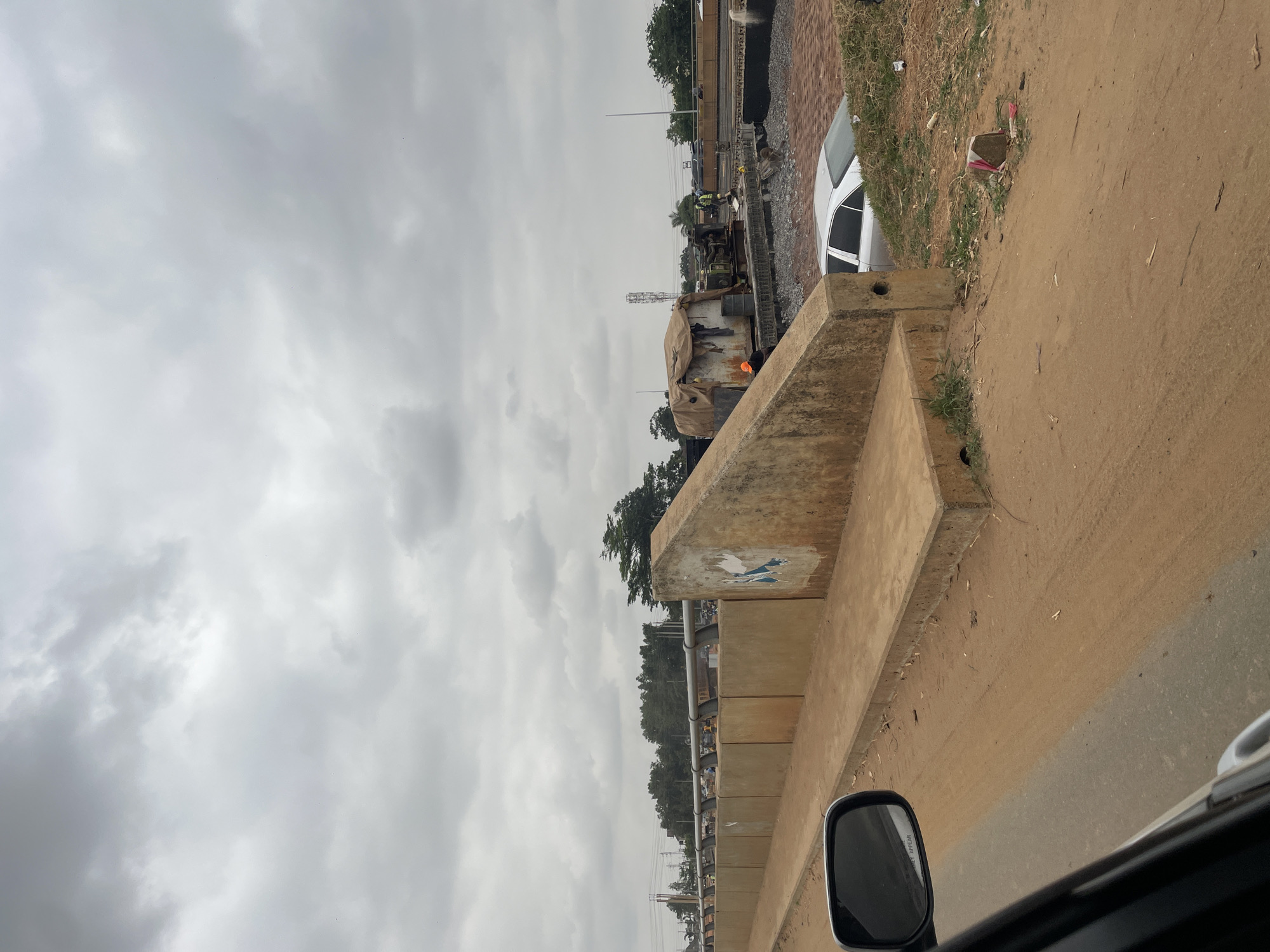 This media file captures an outdoor scene under an overcast, grey sky, likely depicting a construction or infrastructure development area. The image appears to be taken from inside a vehicle, with part of a car's side mirror and roofline visible in the bottom right corner.
In the foreground, a substantial concrete barrier or wall runs horizontally across the lower part of the frame. This barrier shows signs of wear, with patches of faded blue and white paint or graffiti. Adjacent to it, on the right, is a rough dirt shoulder or pathway.
The midground reveals a more active zone. Behind the initial concrete barrier, a white car is parked on what appears to be a gravel or dirt surface. Further left, a long, dark structure, possibly part of a railway track or an elevated road foundation, stretches into the distance. Alongside this structure, there's a bustling work area featuring various construction materials and equipment. A large, dark vehicle, possibly a truck or specialized machinery, is partially covered by a light-colored tarp. A distinctive orange glow is visible near this vehicle, suggesting either an open flame, a light source, or a reflection. Several individuals, likely workers, are present in this area; one person is clearly seen wearing a high-visibility vest. Beyond the immediate construction zone, a line of dense green trees forms the horizon.
The sky above is entirely covered with heavy, diffused grey clouds, indicating an overcast day without direct sunlight. There are no strong shadows, suggesting either an early morning, late afternoon, or a generally dull day. The overall impression is one of ongoing work in a developing landscape. No clearly legible text is visible on any signs or objects, although indistinct markings can be seen on the concrete barrier and a reflection in the car's side mirror is too blurry to decipher. The scene evokes the atmosphere of a busy construction site in an urban or semi-urban setting.