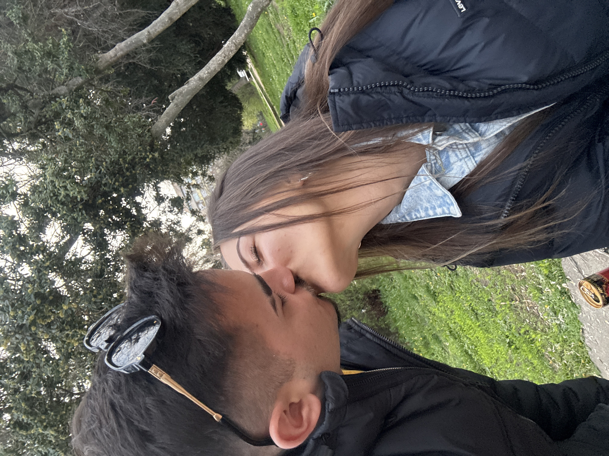 Two people are kissing outside in Sofia, Bulgaria. One individual has sunglasses perched on their head. They are in a grassy area with trees in the background. There is a beverage can nearby. Both individuals are wearing jackets.