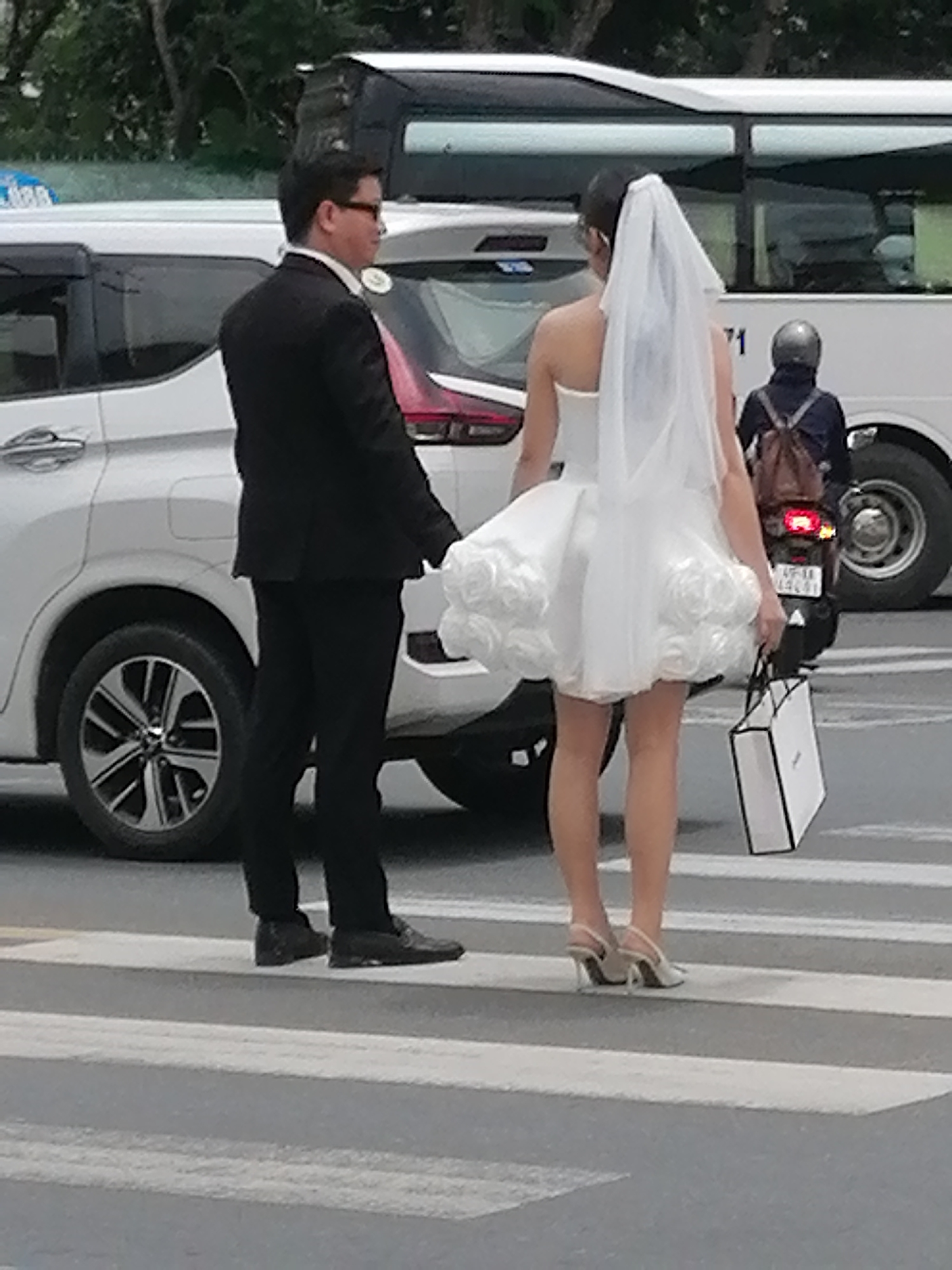In Da Lat, Vietnam, a couple is captured crossing a pedestrian crosswalk. The man, on the left, is dressed in a black suit with a white shirt and dark-rimmed glasses, holding the woman's hand. The woman, on the right, wears a short, strapless white wedding dress with rose embellishments and a veil. She is also carrying a white shopping bag with black trim. In the background, a white bus and a white car are visible, as well as a motorcyclist wearing a helmet and a backpack. The scene appears to be a street in Da Lat, with the characteristic greenery of the city suggested in the background.