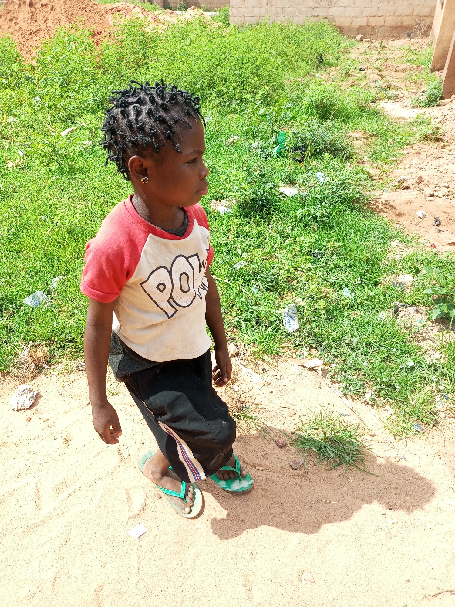 A young child stands in a sandy area next to a patch of green grass and some rubble. The child is wearing a red and white t-shirt with "POW!" printed on it in black letters, black pants, and green flip-flops. The child has their hair braided into small twists and is wearing a gold heart-shaped earring in their left ear. The background shows a pile of reddish dirt and a brick wall.