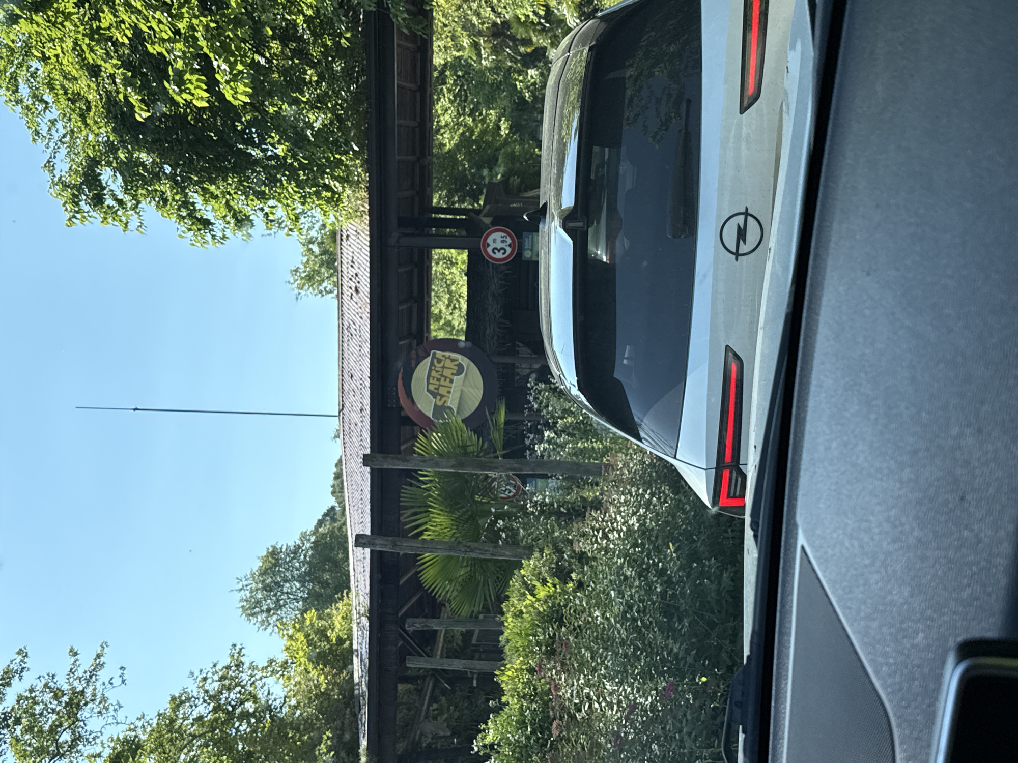 The image depicts the entrance of the Parco Natura Viva in Bussolengo, Italy, as seen from inside a car. The park entrance is marked by a wooden structure with signage. A white Opel automobile is visible partially obstructing the view of the entrance. The environment includes greenery and trees typical of a park setting. A height restriction sign is also visible on the structure.