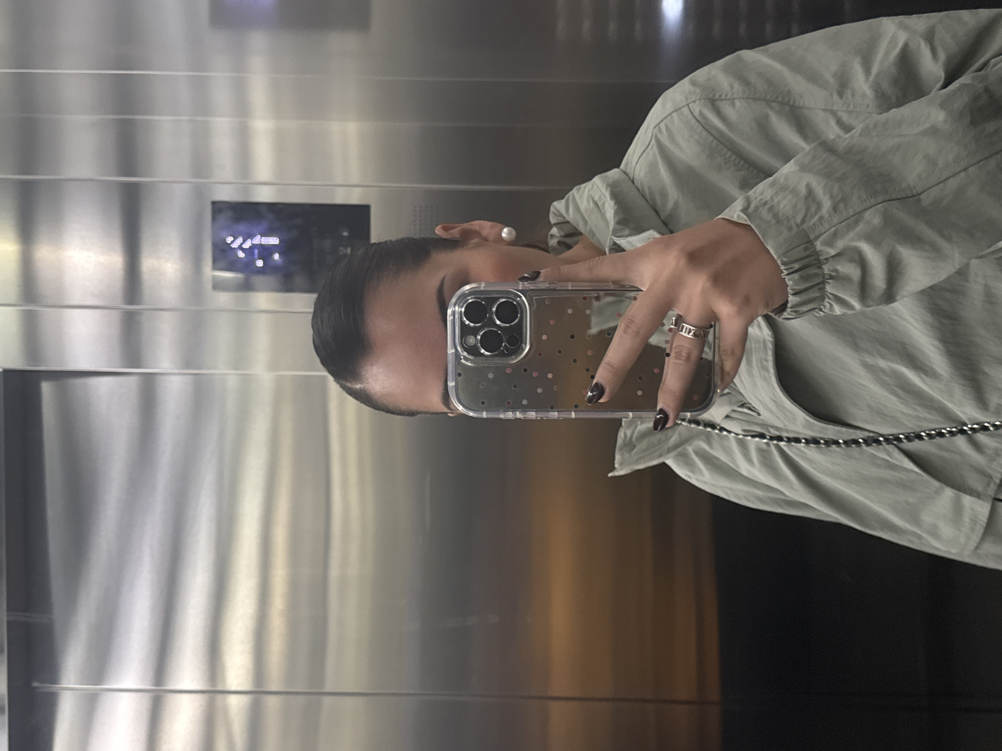 A person is seen taking a mirror selfie inside an elevator. The image is oriented vertically, showing the person from the chest up, holding a smartphone to capture their reflection.
The individual, whose face is mostly obscured by the phone, has dark hair pulled back and wears a single pearl stud earring on their right ear. They are dressed in a light olive green or grey lightweight jacket. On their left hand, holding the phone, they wear a silver-toned ring on the ring finger, featuring Roman numerals, specifically "IIX" and "I" are discernible. Their fingernails are painted with a dark, possibly burgundy or brown, polish.
The smartphone is an iPhone model with a triple-lens camera system, encased in a clear cover adorned with small, colorful polka dots. A silver-toned chain, possibly a phone strap or part of an accessory worn under the jacket, is visible hanging from the phone or wrist area.
The background consists of the brushed metallic walls of an elevator interior. To the left of the person's head, an illuminated control panel is visible, displaying blurry, light blue characters that appear to be floor indicators or operational symbols, although they are not legible.
The activity depicted is a mirror selfie taken within an elevator, likely in San Bernardino Tlaxcalancingo, Mexico, as per the provided location context. The lighting suggests an indoor setting, typical of an elevator.