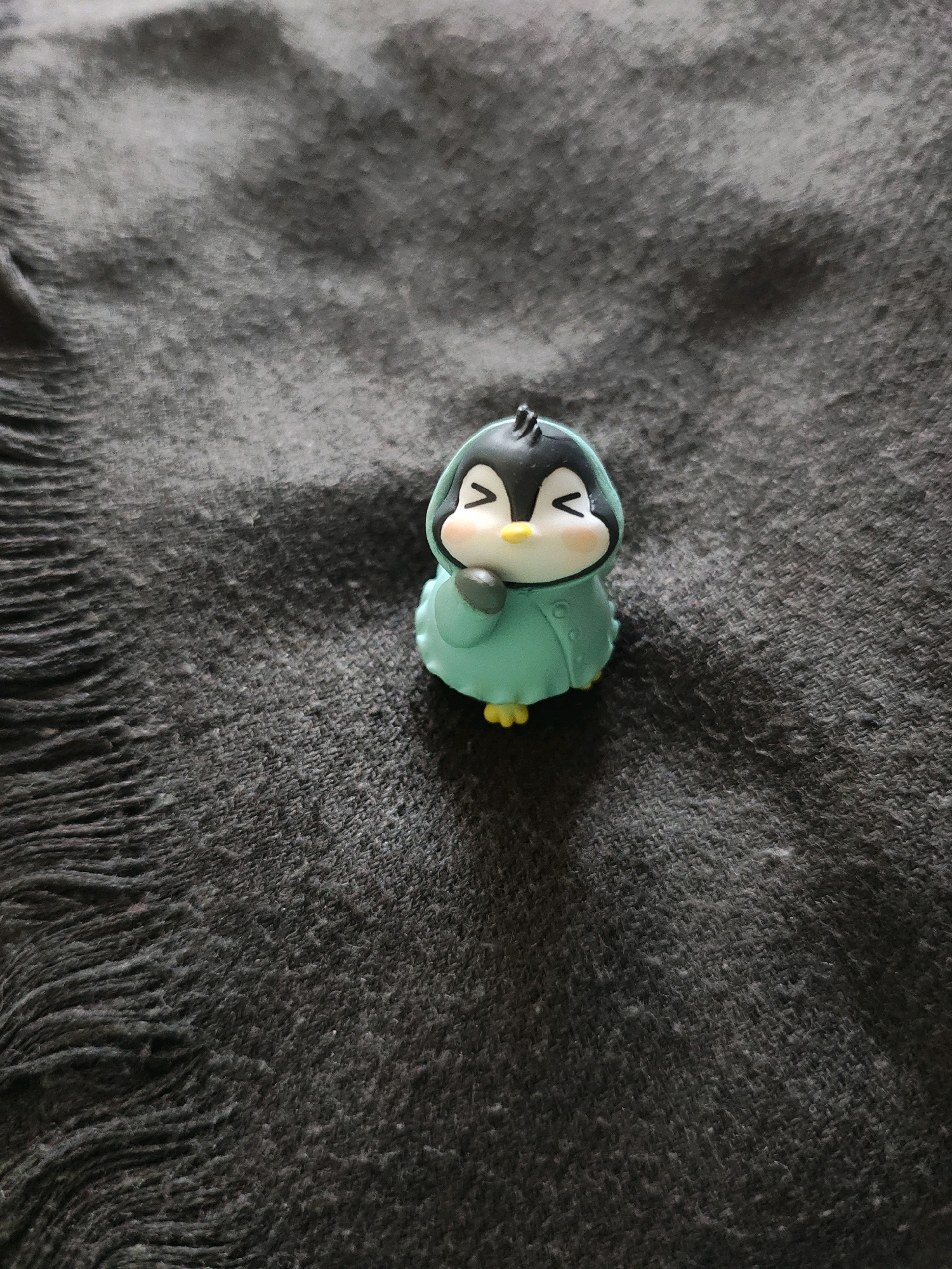 A miniature figurine of a penguin, wearing a turquoise hooded coat and yellow feet, sits on a dark, textured fabric. The penguin's eyes are closed and it has rosy cheeks, giving it a cheerful expression. The fabric has a fringe on the left side. The scene is lit from the top right, casting a shadow to the bottom left of the figurine. The location within Jaipur Municipal Corporation, India, is not discernible from the image.