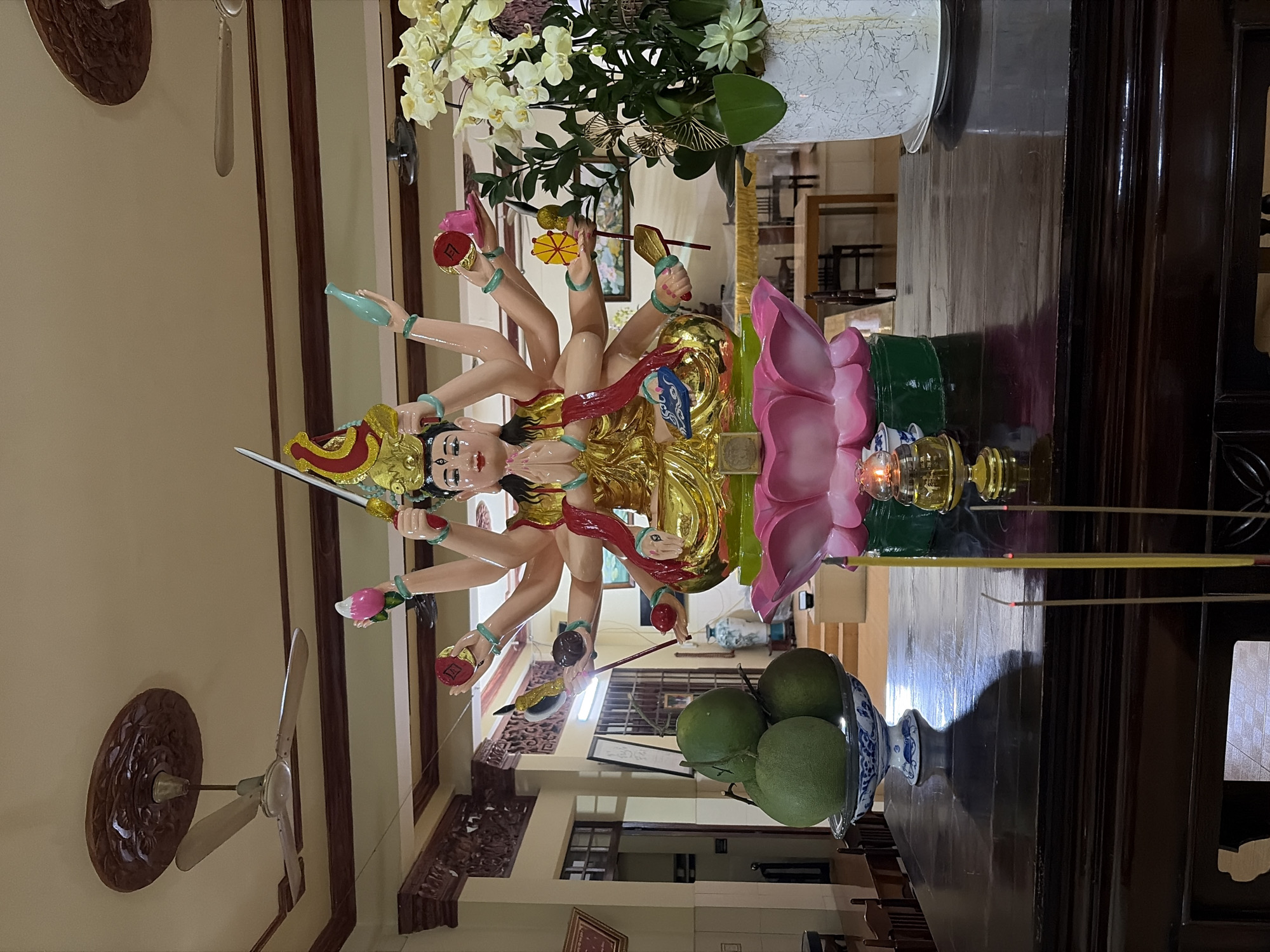 The image shows an altar within a building, likely a temple or shrine, in Thủ Đức, Vietnam. At the center of the altar is a multi-armed statue, possibly of a deity, adorned in gold and seated on a pink lotus. The statue holds various symbolic objects in its many hands, including a sword and what appears to be a lotus flower.
In front of the statue, on a dark wooden surface, are several items associated with worship: a bowl filled with green fruits, likely pomelos, a small oil lamp with a lit flame, and several lit incense sticks emitting smoke. To the right of the statue, a large white pot contains a bouquet of light-colored orchids.
The background reveals architectural details of the building, including ornate wooden carvings on the ceiling and walls. Two ceiling fans are visible, one on the left and one on the right side of the frame. Further back, doorways and interior spaces of the building can be seen, suggesting a communal or religious setting. The overall impression is one of a serene and devotional space.