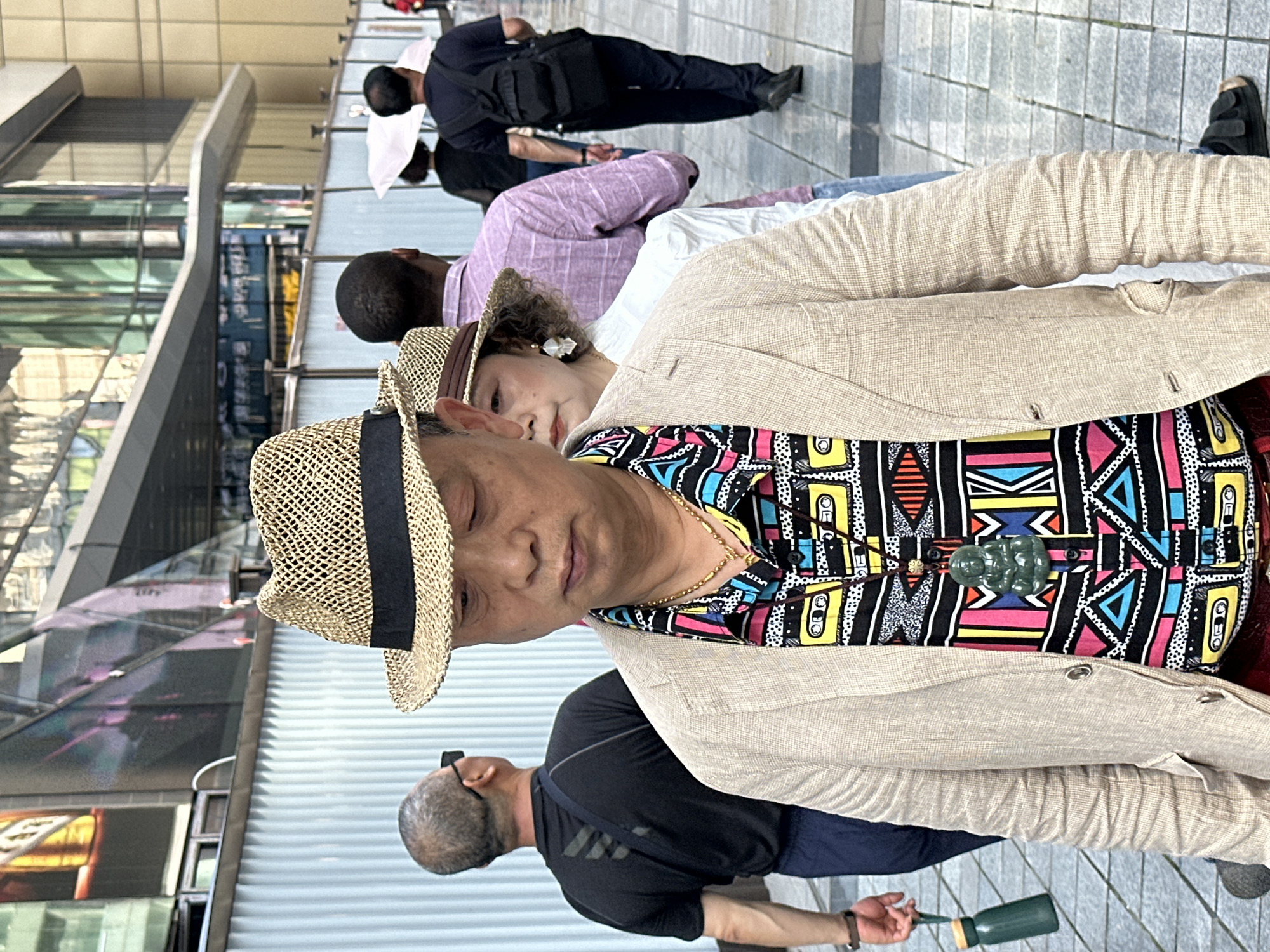 The image depicts a group of people walking in an urban environment, possibly in the Yuzhong District of China. A man in the foreground is wearing a straw hat with a black band, a colorful patterned shirt, and a light brown blazer. He has a gold chain and a green pendant. To his left, a woman with curly hair and a white flower accessory is partially visible. In the background, other people are walking, including a man in a black shirt and a man in a lilac shirt. The scene appears to be on a paved walkway with buildings in the background. A bottle, possibly green glass, is visible in the lower part of the frame. The image is slightly angled.