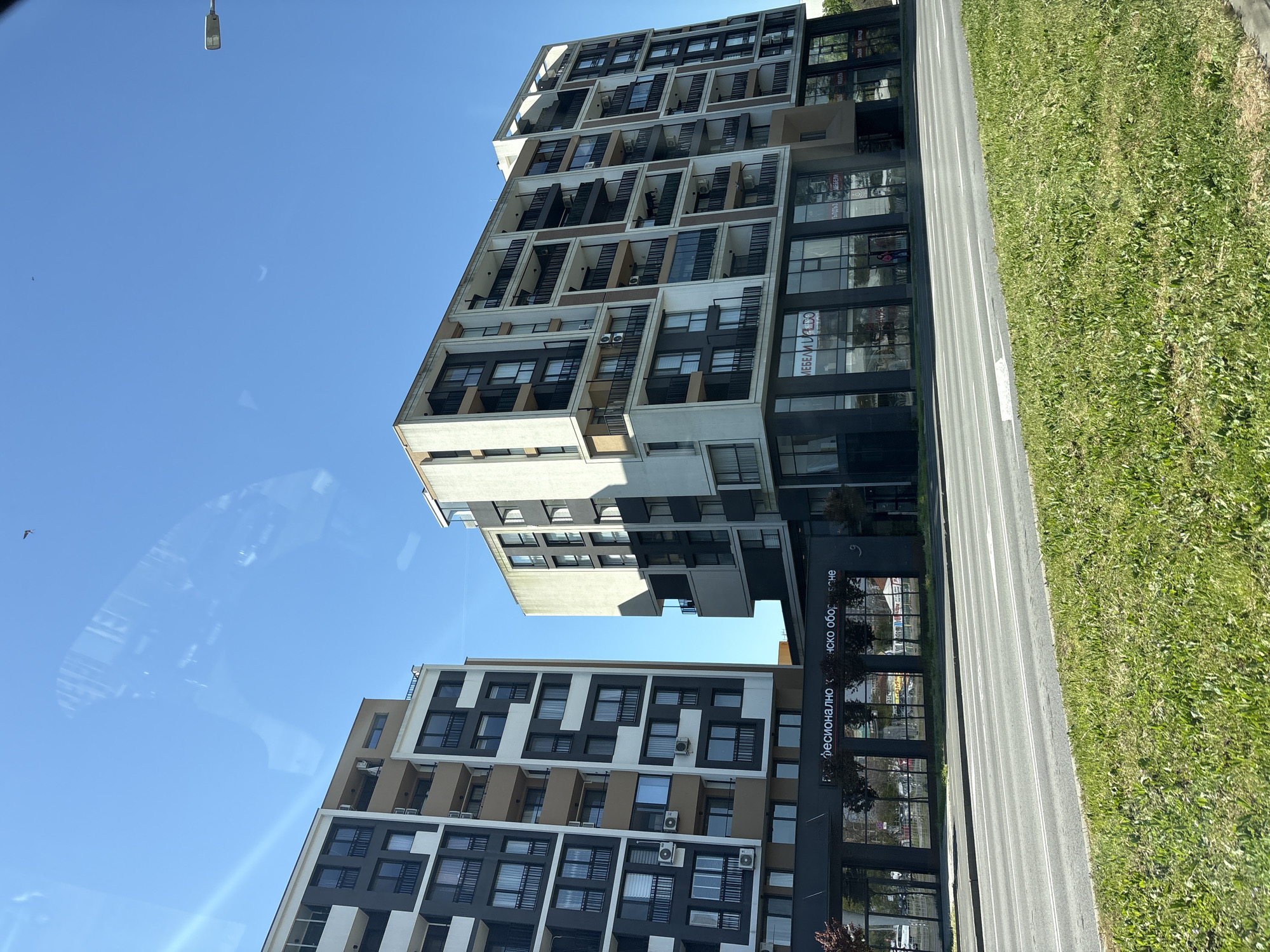 Here is a description of the image:
The image shows two tall, modern apartment buildings next to a road and grassy area. The buildings feature a mix of white, gray, and dark brown facades with numerous windows and balconies. The sky is bright blue and clear. A street light is visible at the top of the frame. The image is taken from a low angle, suggesting the viewpoint of someone standing or driving nearby. Along the building's base, shop windows are visible, with a sign in a language other than English.