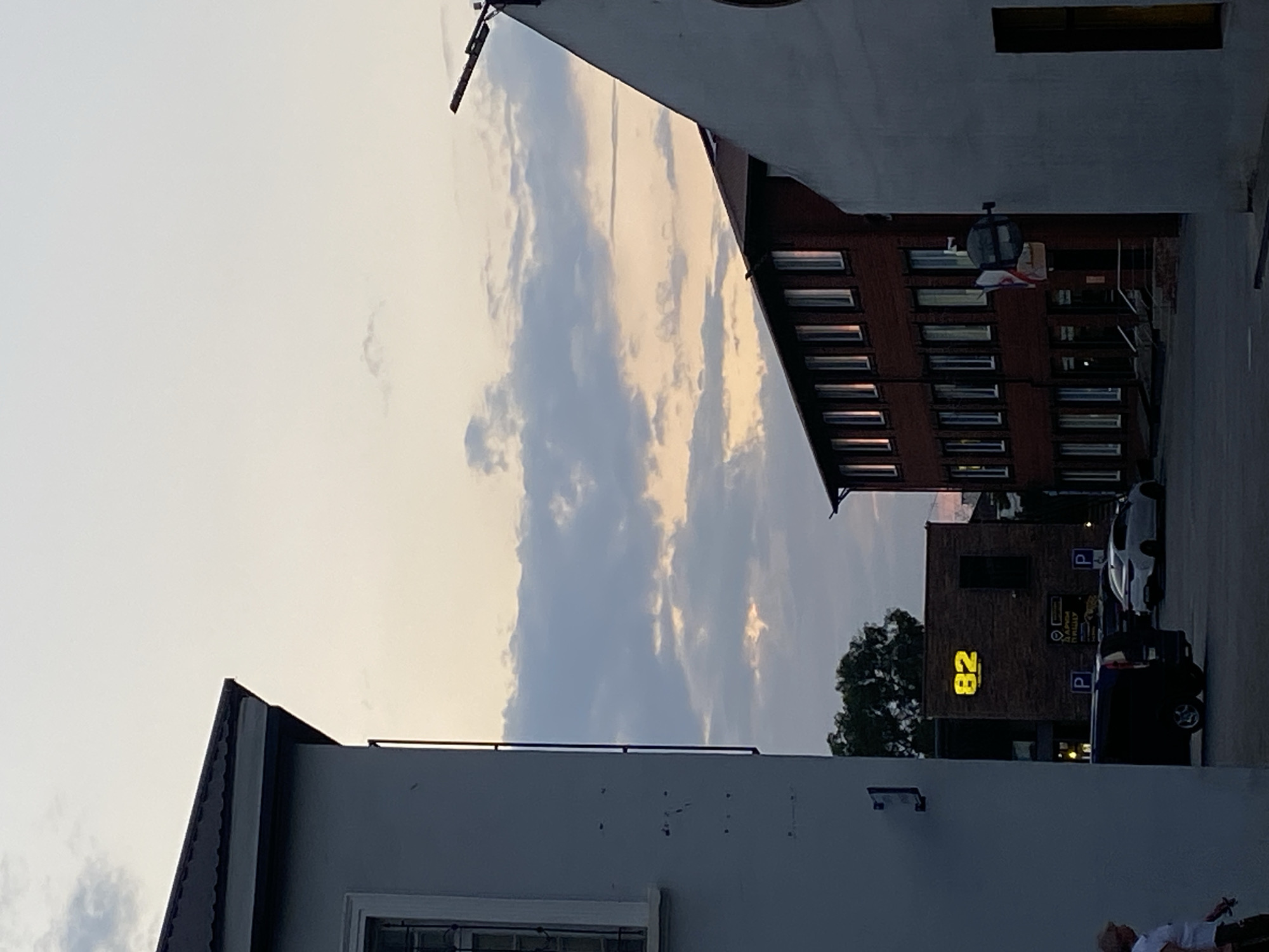 The image shows a street scene at dusk in Feodosia, Ukraine. In the foreground, a light-colored building occupies the left side of the frame. To the right, a dark red brick building with multiple windows runs diagonally into the background. The sky above is a pale gradient of blue and pink, with scattered clouds.
In the middle ground, a dark-colored van is parked on the street. Further back, a dark brick building has large, illuminated yellow numbers "82" on its facade. Two parking signs with the letter "P" are visible near the street. A white car is parked on the right side of the street.
A person is partially visible on the far left, walking away from the camera. Another person is seen on the far left edge of the image, partially obscured by the building. A French flag is mounted on the facade of the red brick building. The scene appears to be a commercial or residential area, with elements of urban infrastructure.