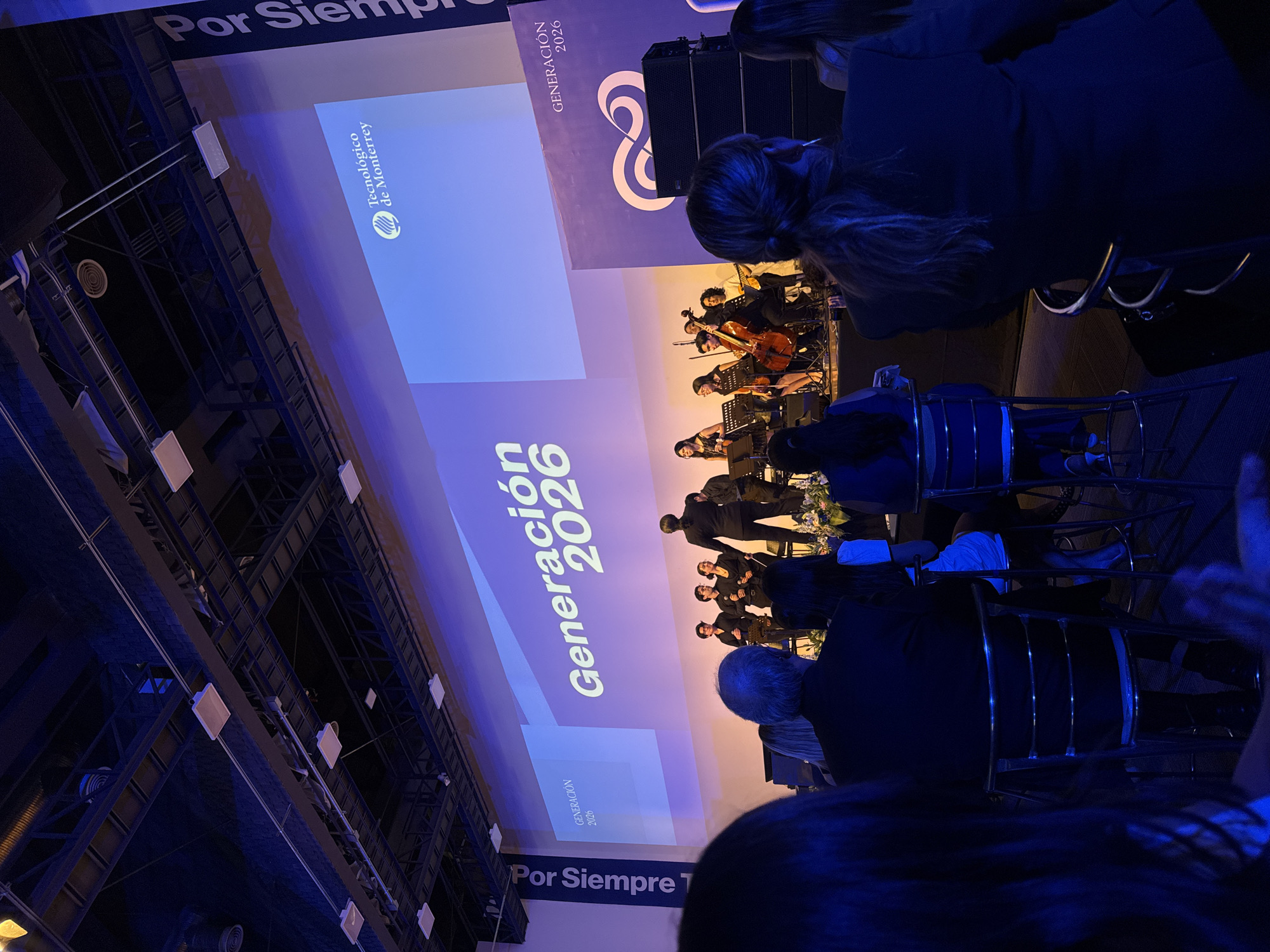 The image depicts a formal event held at the Tecnológico de Monterrey campus in Puebla, Mexico. The focus is on a stage presentation with a screen displaying the text "Generación 2026" and the Tecnológico de Monterrey logo. A musical ensemble is performing on stage, visible on the screen. The audience is seated in rows of metal chairs, with attendees dressed in formal attire. Overhead, there is a network of structural supports with visible lighting and ventilation fixtures. The scene is lit with blue lighting, creating a formal and focused atmosphere.