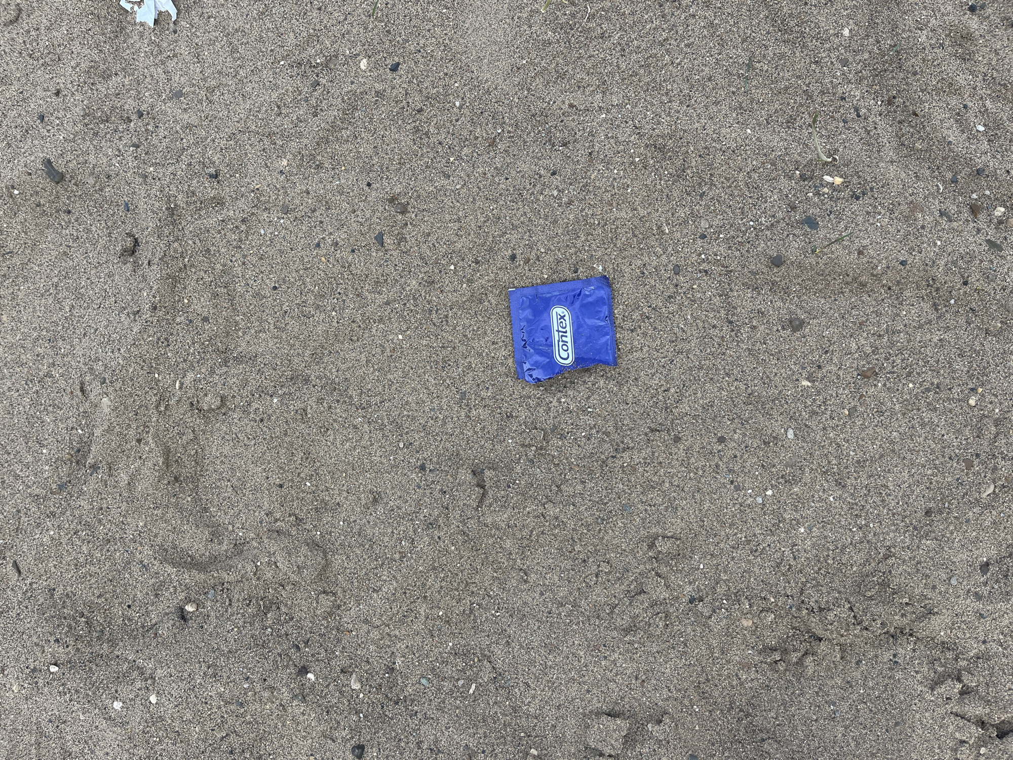 The image shows a single, blue condom wrapper on a sandy surface. The wrapper has the brand name "Contex" printed in white. The sandy surface appears to be outdoors, with small pebbles and debris scattered throughout. The lighting suggests it is daytime. The location is identified as the bank of the Kama River in Chaikovsky, Russia, a recreational area often frequented by people.