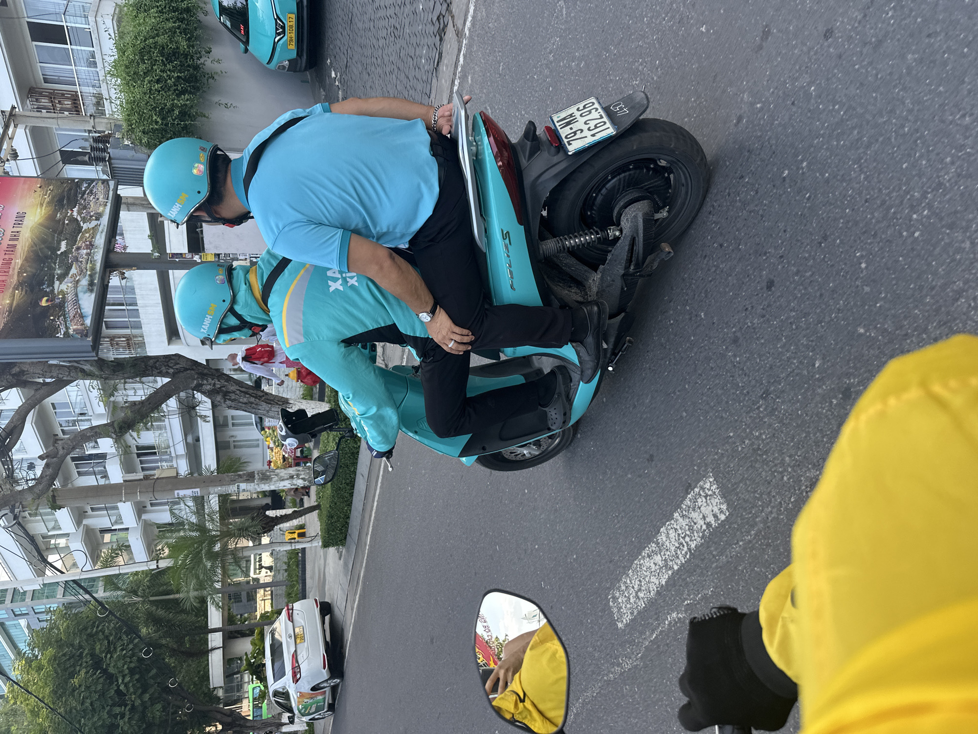 The image provides a dynamic street-level view from the perspective of a motorcycle rider, featuring a bright yellow jacket and a gloved hand on the handlebars in the foreground. Reflected in the motorcycle mirror is the person in the yellow jacket.
The central focus is on two individuals riding a light blue/turquoise scooter, likely part of a ride-hailing or delivery service. Both are clad in matching light blue/turquoise helmets, prominently displaying the "XANH SM" logo, and similar colored uniforms. The scooter's brand, "FELIZS," is visible on its side, and its license plate, "79-NA 162.96," is clearly displayed at the rear. The passenger, seated behind the driver, wears a short-sleeved shirt and black trousers, while the driver wears a long-sleeved shirt.
The scene unfolds on an urban street lined with multi-story buildings, interspersed with lush trees and utility poles. A white car is parked along the curb, and further ahead, a light blue taxi, also branded "XANH SM" and bearing the license plate "79H-108.17," is visible. A prominent billboard on the left side features text including "NHA TRANG" and "DA TRUNG TÂM NHA TRANG," strongly indicating the location as Nha Trang, Vietnam. The road surface is asphalt with white painted markings. The image suggests a bright, clear daytime setting, capturing the typical activity of a Vietnamese city street.