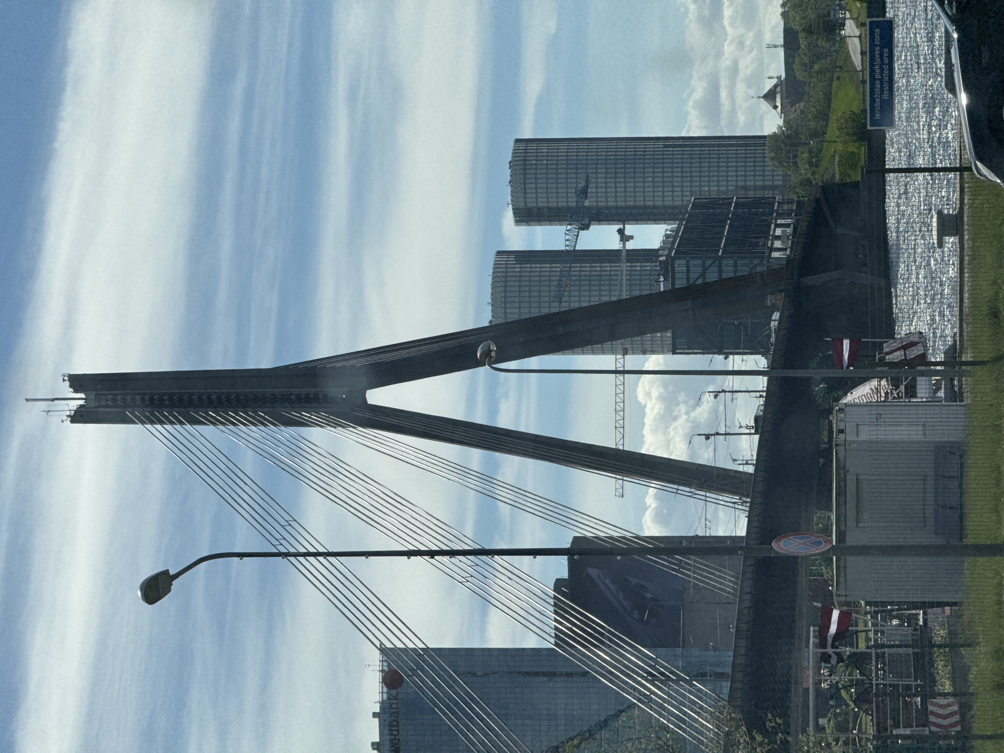 The image depicts the Vansu Bridge in Riga, Latvia, a cable-stayed bridge spanning the Daugava River. The bridge's distinctive A-shaped pylon is prominent against a sky with streaky clouds. Modern buildings, possibly offices or apartments, are visible in the background. A construction crane is situated between the buildings. In the foreground, a streetlight is seen near the bridge's support cables. There's a sign indicating a restricted area in Latvian and English. A Latvian flag is visible, partially obscured. A small church spire is visible in the far distance. There are various objects in the foreground including road signs, fencing, construction materials, and a container. A part of a vehicle is visible on the right-hand side of the image.