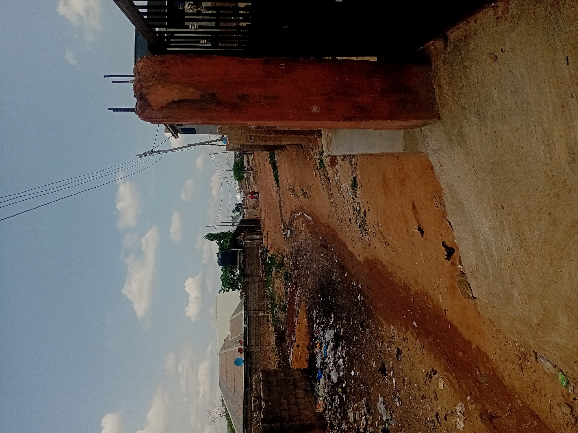 The image, rotated 90 degrees clockwise, presents an outdoor scene dominated by an unpaved, reddish-brown dirt path under a light blue sky with scattered white clouds. In the foreground, a large, textured orange-red concrete or plastered structure, possibly a pillar or wall, is prominent, with several reinforcing bars (rebar) protruding from its top edge. Above this structure, a dark metal gate or railing is partially visible.
The dirt path extends into the distance, appearing dusty and uneven. Along the right side of the path, particularly in the middle ground, there is a considerable accumulation of scattered rubbish and debris. Further down the path, a lone individual wearing red is visible walking or standing.
Power lines stretch across the mid-ground, supported by utility poles that line the left side of the path. In the middle distance, various structures are present, including buildings with corrugated roofs and walls constructed from cinder blocks. A black water tank stands on a support structure, and two balloons (one blue, one reddish) are tied to a fence-like element. The scene suggests a semi-urban or developing area, possibly in a warm climate, characterized by basic infrastructure and open ground. No discernible text is visible.