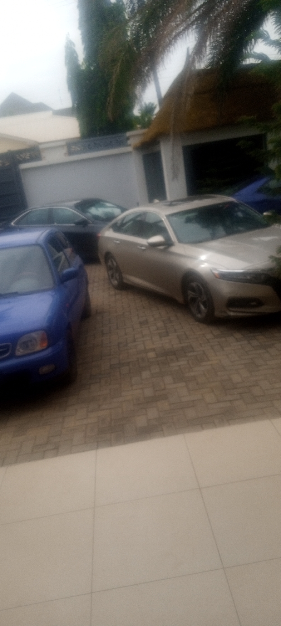 The image shows a driveway with three cars parked in front of a building. The car closest to the viewer is blue, followed by a black car and a gold car. The driveway is paved with rectangular stones. The building in the background has a white wall with a black metal gate. There is also a small hut with a thatched roof. The location is potentially Unguwar Rimi, Nigeria, based on the user's specification. No people are visible in the image. The scene appears to be a residential setting.