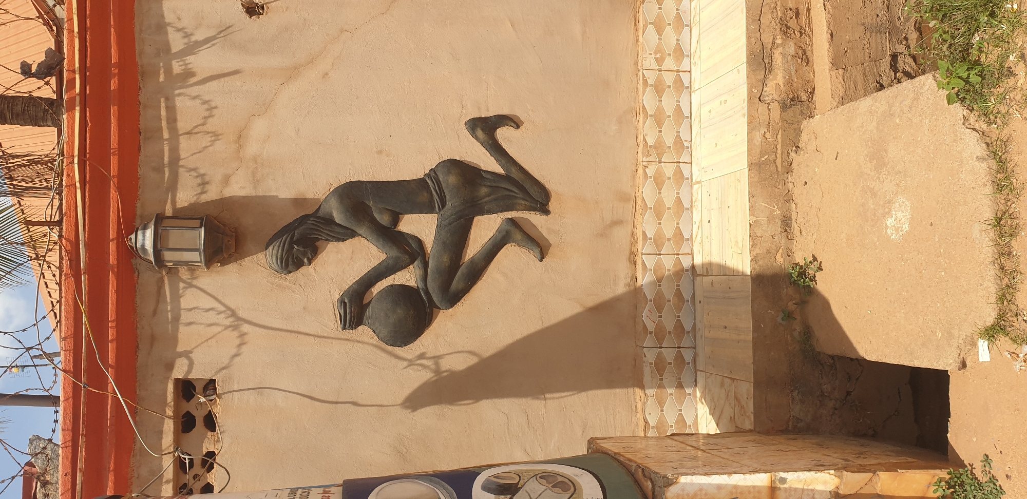 A bas-relief sculpture of a woman with a pot is affixed to a textured, beige wall in Sabon Tasha, Nigeria. The sculpture depicts a woman with her hair braided, wearing a draped cloth around her waist, kneeling on her right leg with her left leg bent. She is holding a round pot with both hands. To the left of the sculpture, a metal lantern is mounted on the wall. Above the wall, barbed wire and palm fronds are visible against a bright sky. Below the wall, a tiled border runs horizontally, and a set of concrete steps descends into a shallow, dirt-filled pit. The overall scene suggests an outdoor setting, possibly a courtyard or entrance.