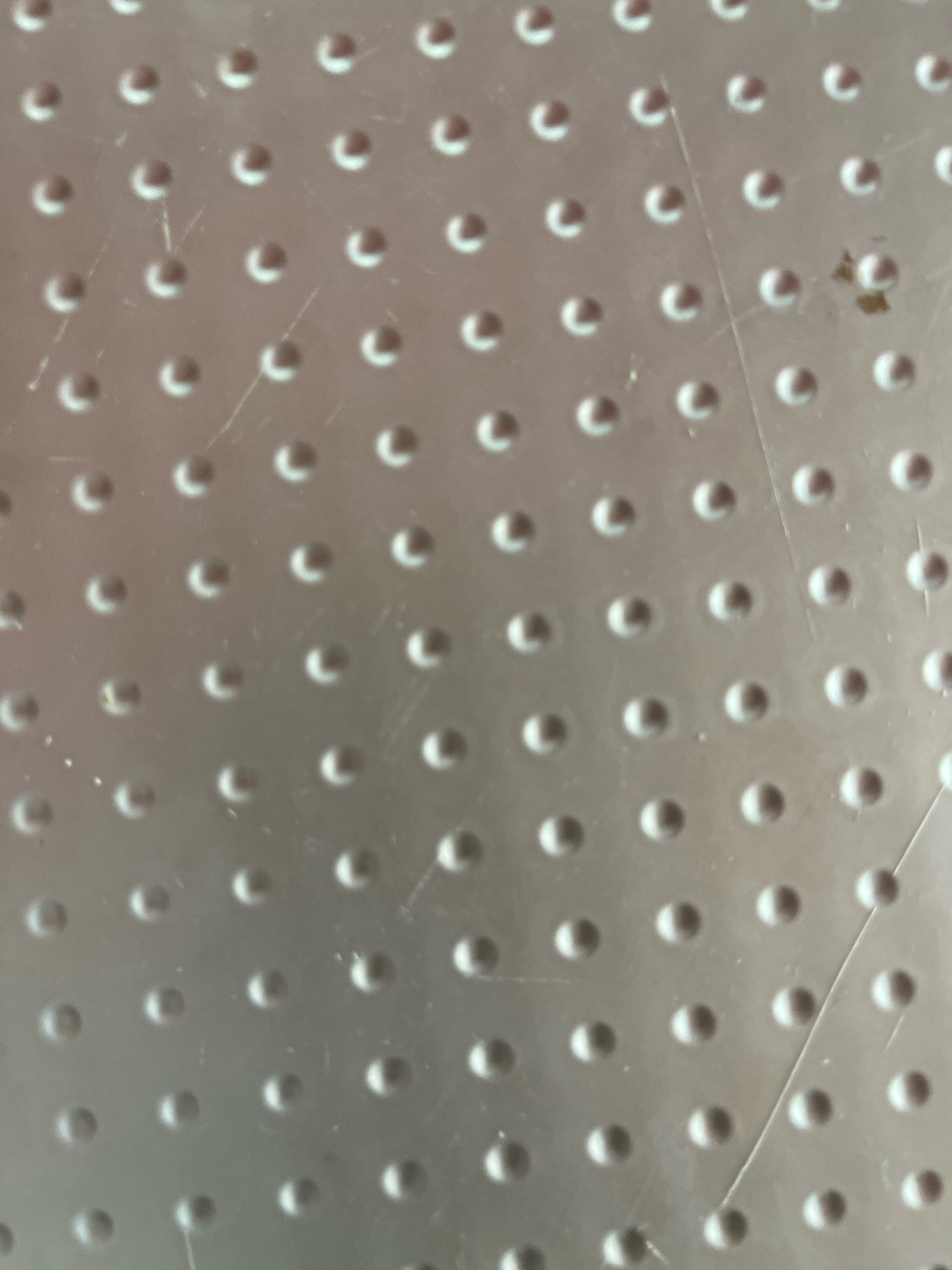 The image displays a close-up, overhead view of a textured metallic surface characterized by a repeating pattern of small, raised circular dots. The surface appears to be a light, silvery-gray color and shows some minor scratches and a dark speck of debris in the upper right quadrant. The lighting creates subtle shadows around the dots, emphasizing their three-dimensional form. The scene is devoid of any context that would place it in Gaushala, Nepal or any other specific location.