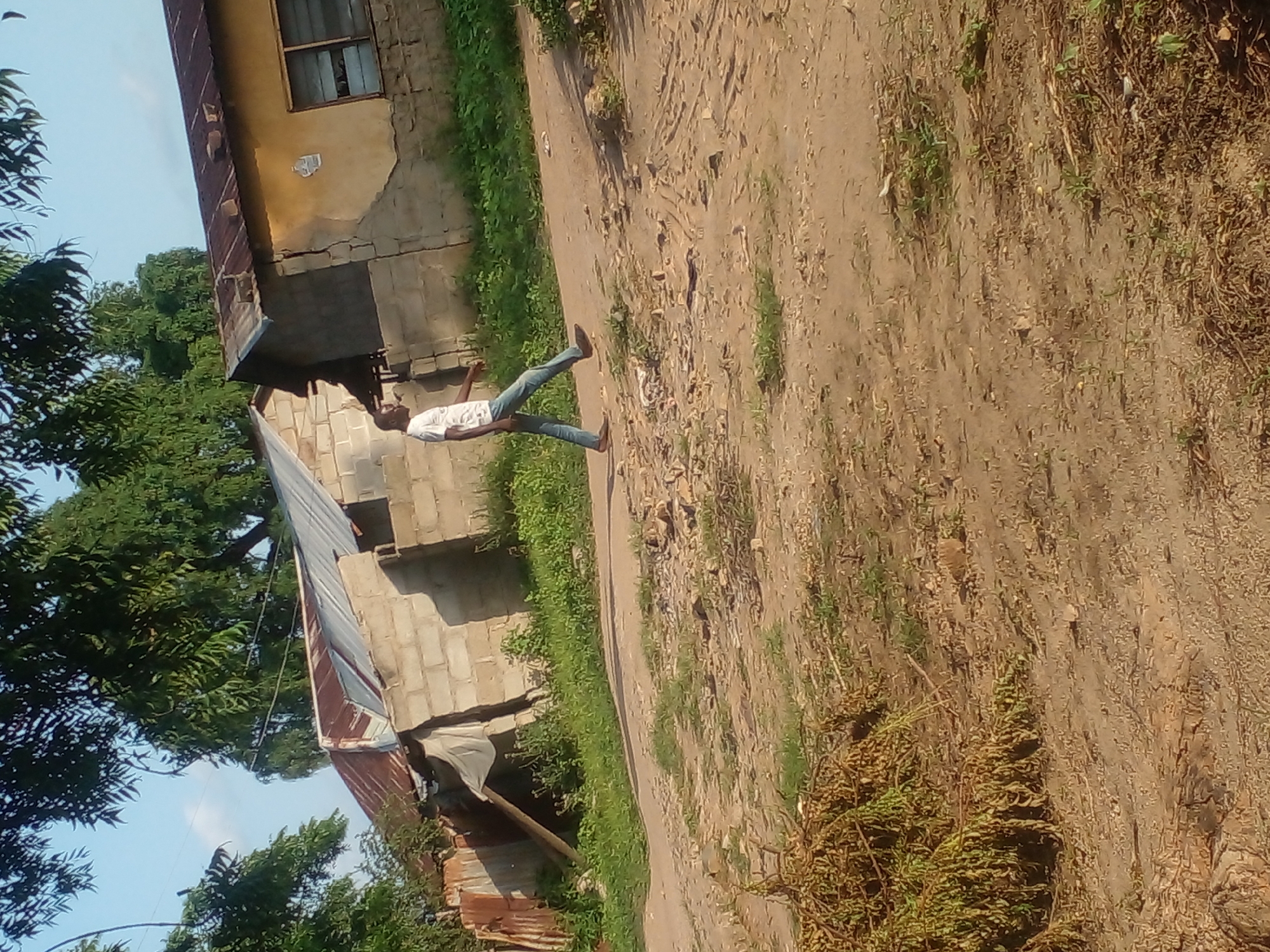 In the city of Langtang, Nigeria, a man walks down a dirt road. He is wearing a white t-shirt and blue jeans. The road is surrounded by dry grass and scattered rocks. In the background, there are several unfinished buildings with corrugated metal roofs. The sky is clear and blue.